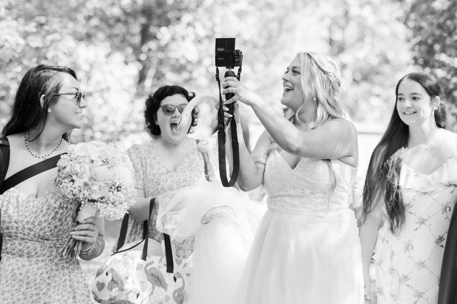Bride in white gown laughs while holding camera with bridesmaids outdoors in black and white wedding photo in Airlie Gardens in Wilmington,