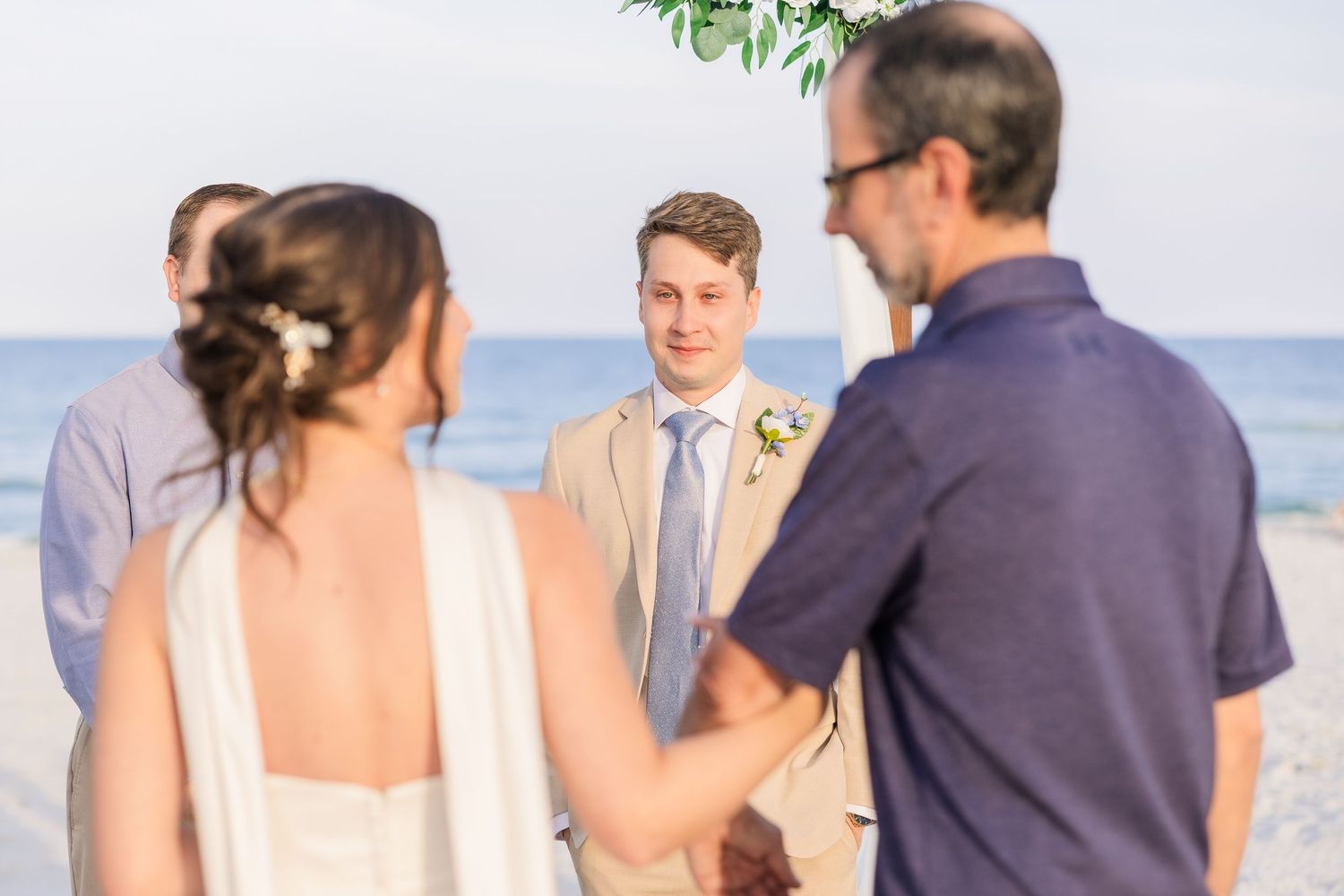 Groom in tan suit smiles at bride during intimate beach wedding ceremony with ocean backdrop in Carolina Beach near Wilmington, NC.