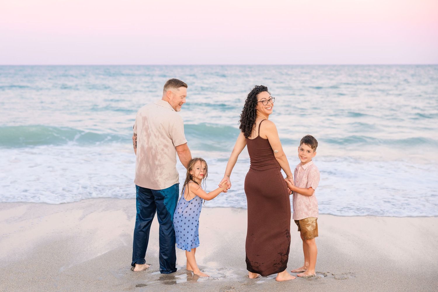Family of four holding hands on beach at pink sunset by ocean.
