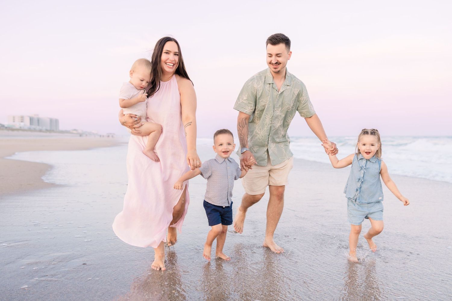 Happy family of five walking barefoot on a sandy beach at sunset, parents holding hands with young children in pastel outfits.