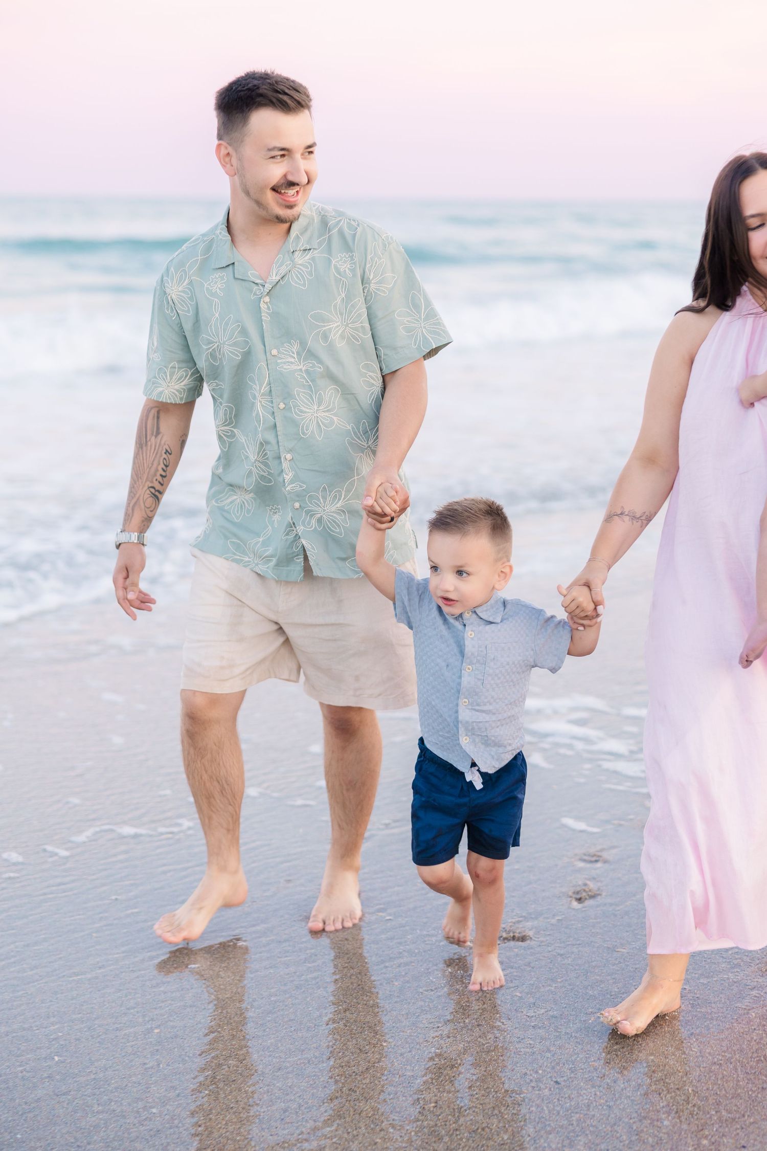 Young parents walk barefoot on beach at sunset holding hands with their toddler son in blue outfit.