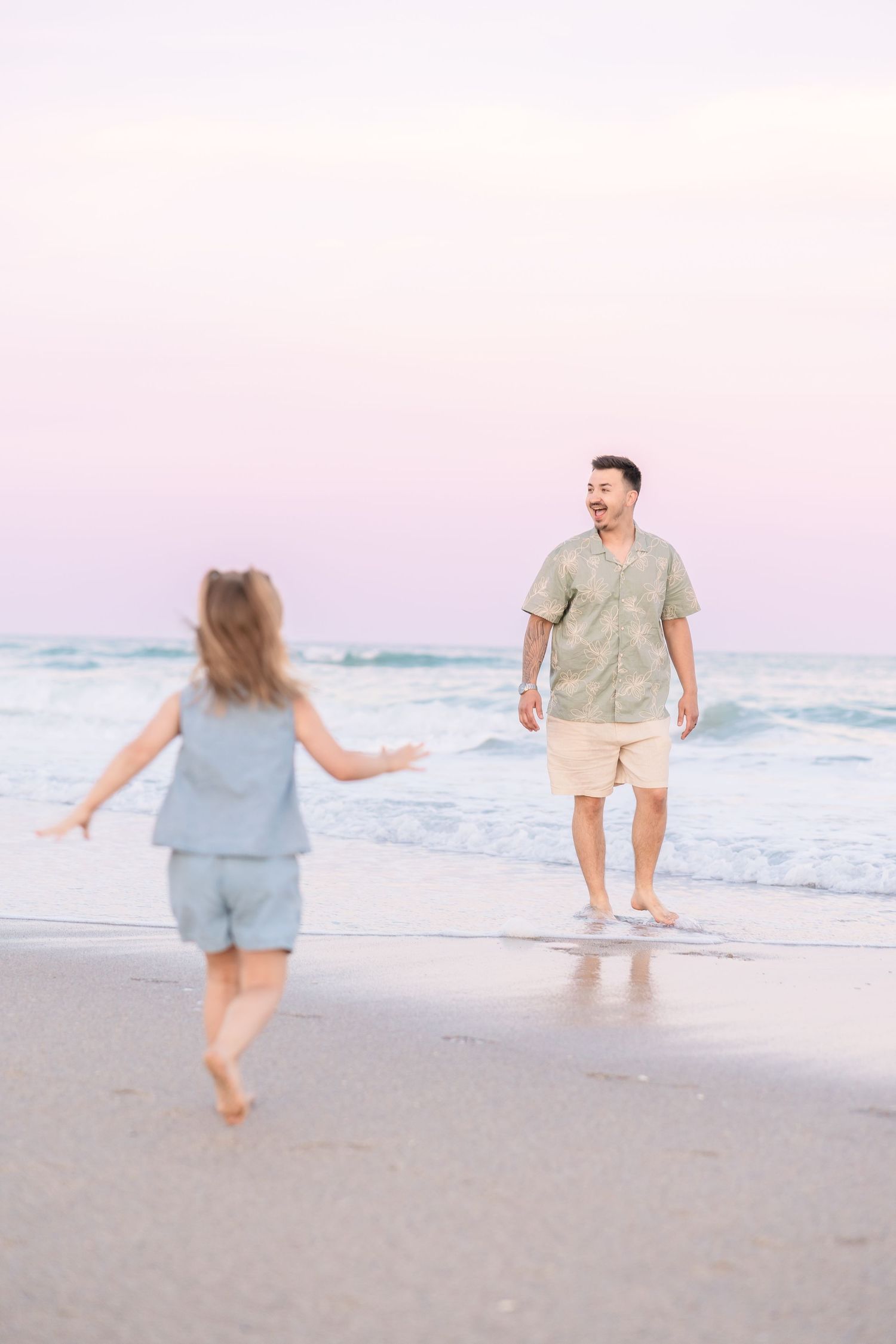 Laughing father opens arms as young girl in blue outfit runs toward him on sunset beach.