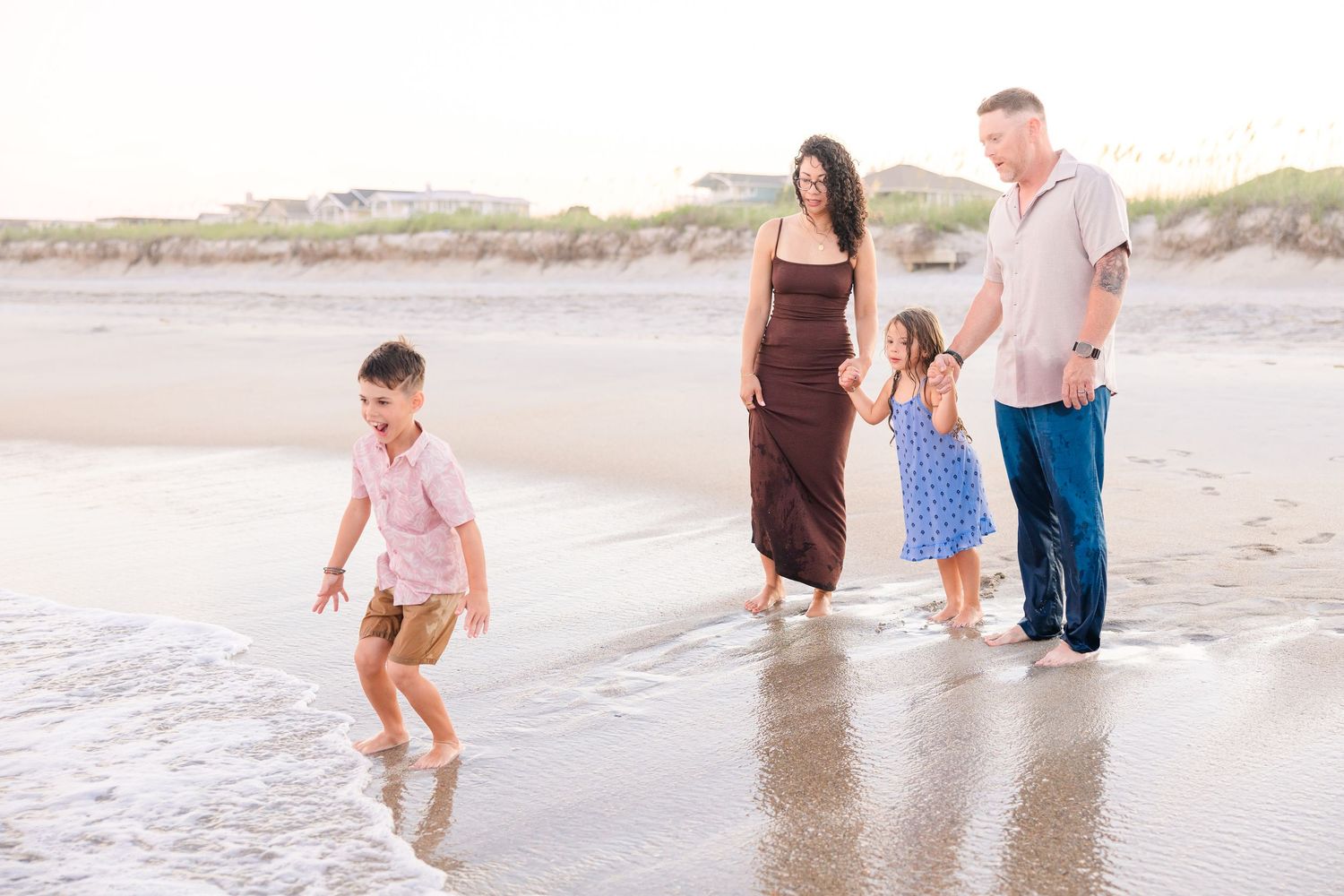 Family of four walks along shoreline at sunset, with young boy running toward gentle ocean waves.