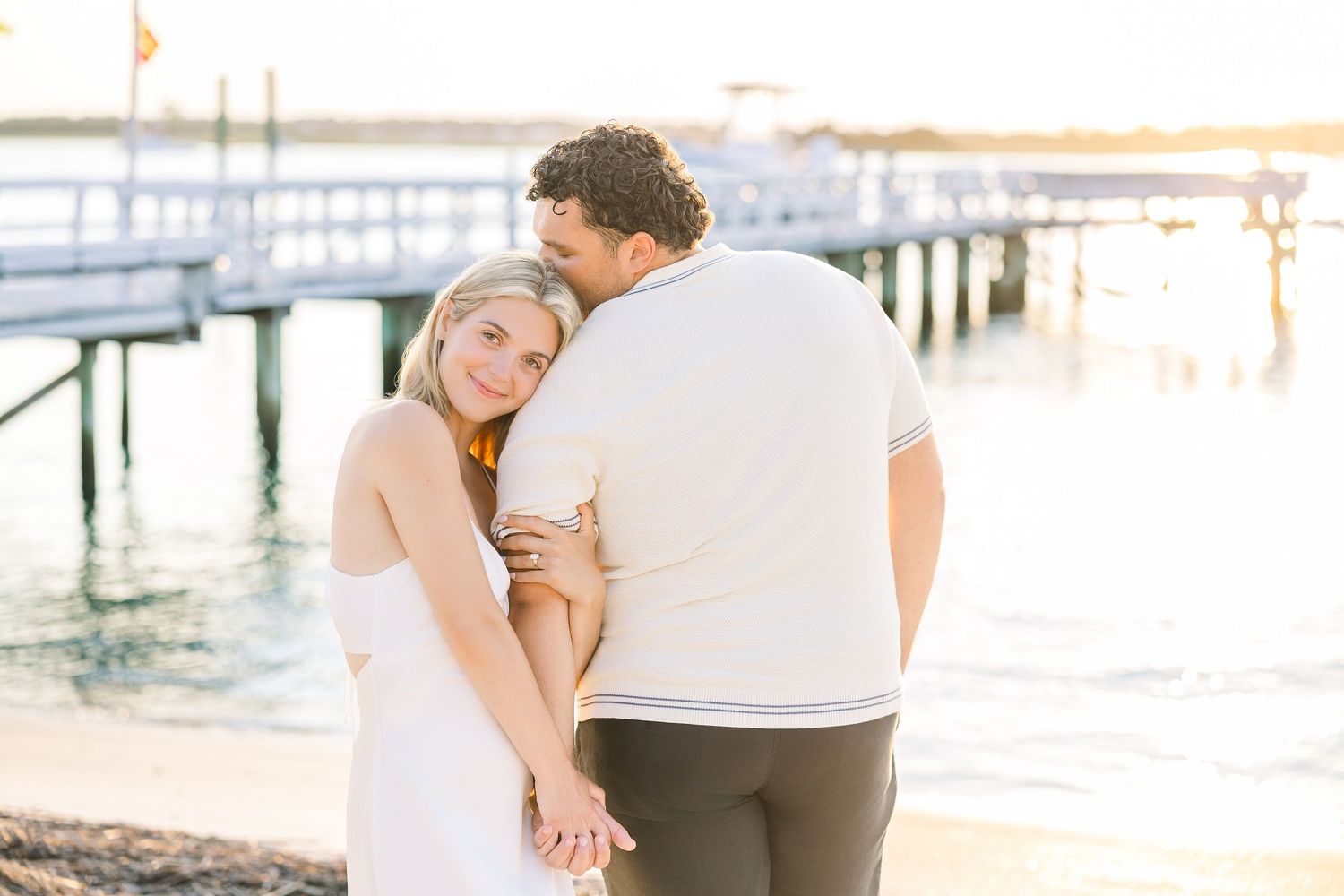 Couple embracing near waterfront pier at golden hour, woman in white dress holding hands with partner.