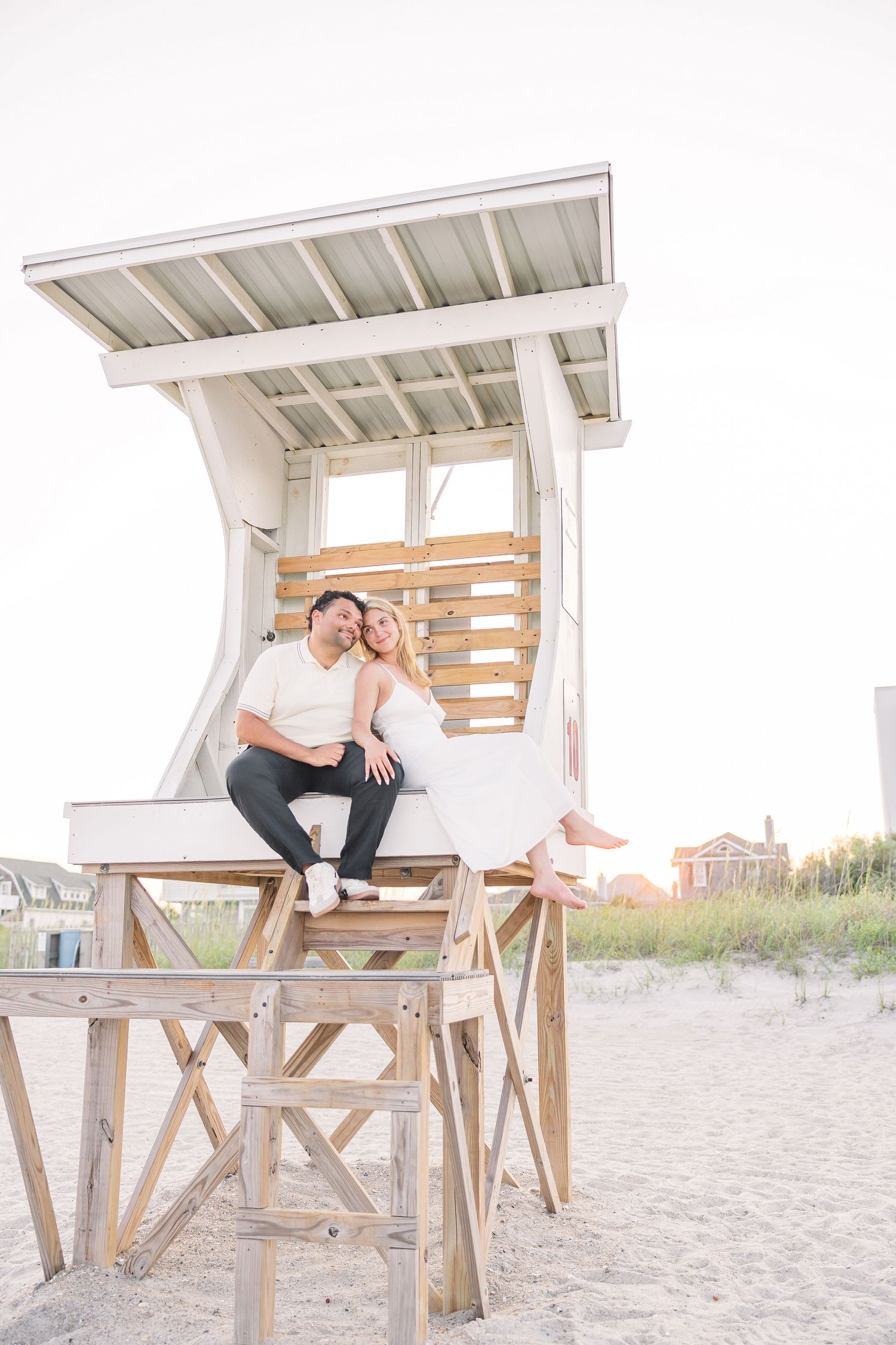 Couple sitting together on a white lifeguard tower on a sandy beach at golden hour.