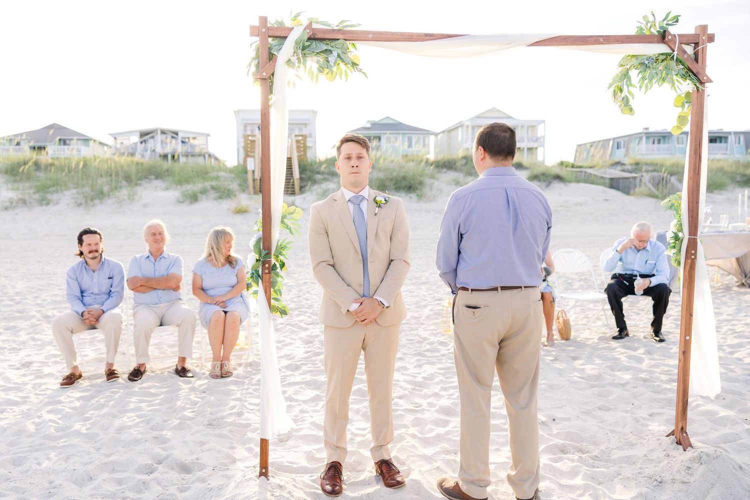 Groom stands at rustic wooden beach wedding arch waiting for bride as small group of guests are seated in Carolina Beach near Wilmington, NC