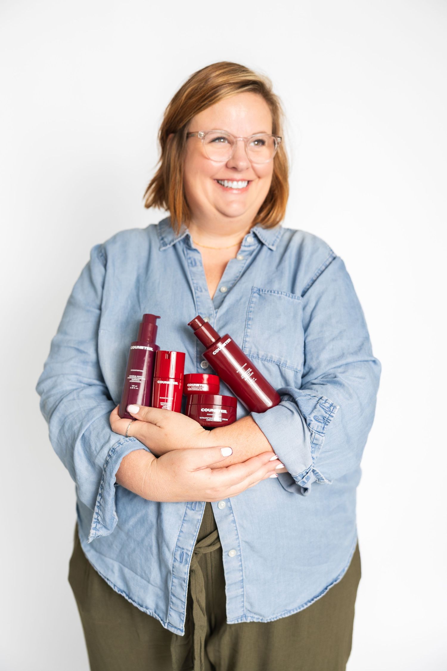 Person in denim shirt holding multiple red cosmetic products against white background.