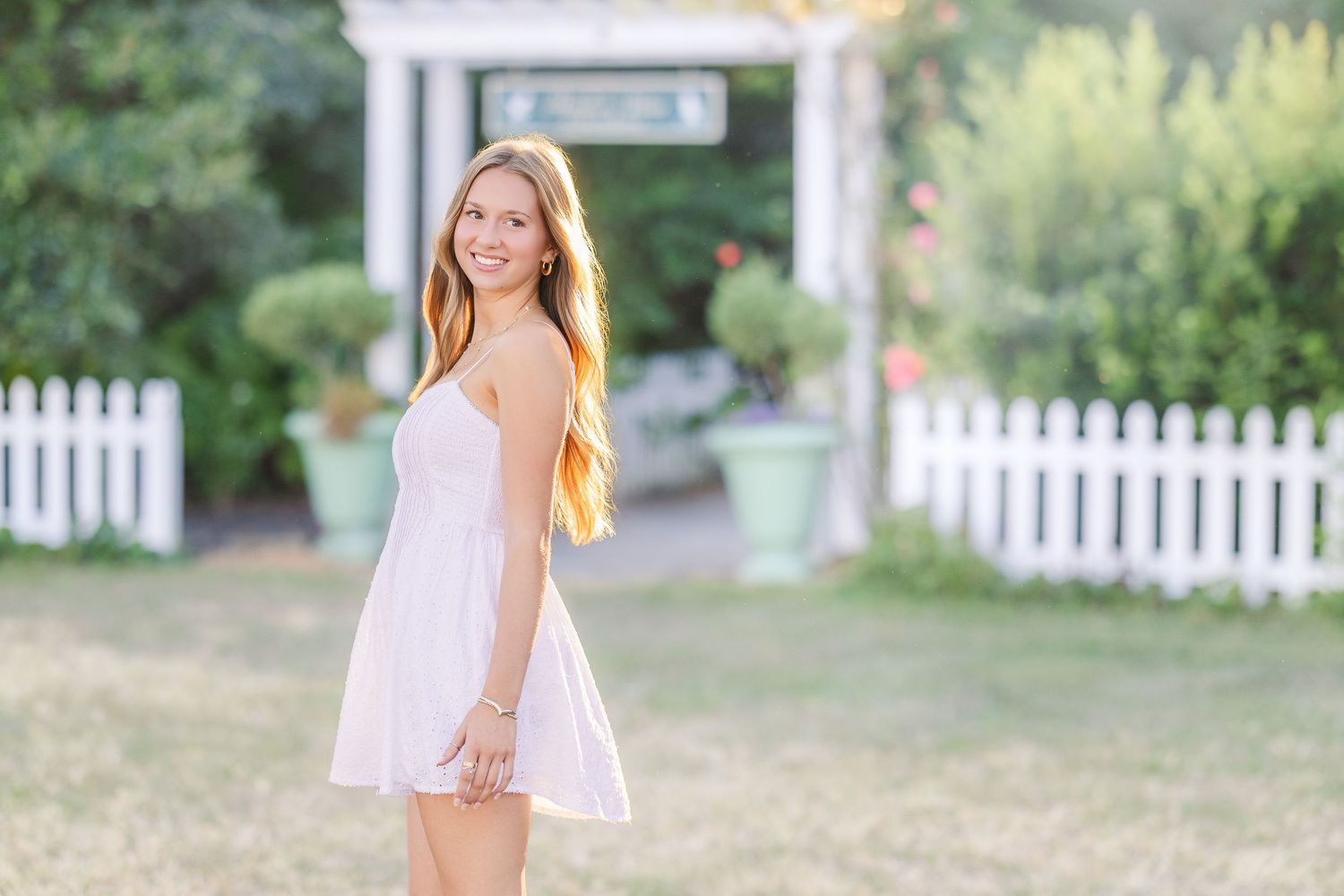 Someone in a breezy white dress walks along a garden path with white fence in background.