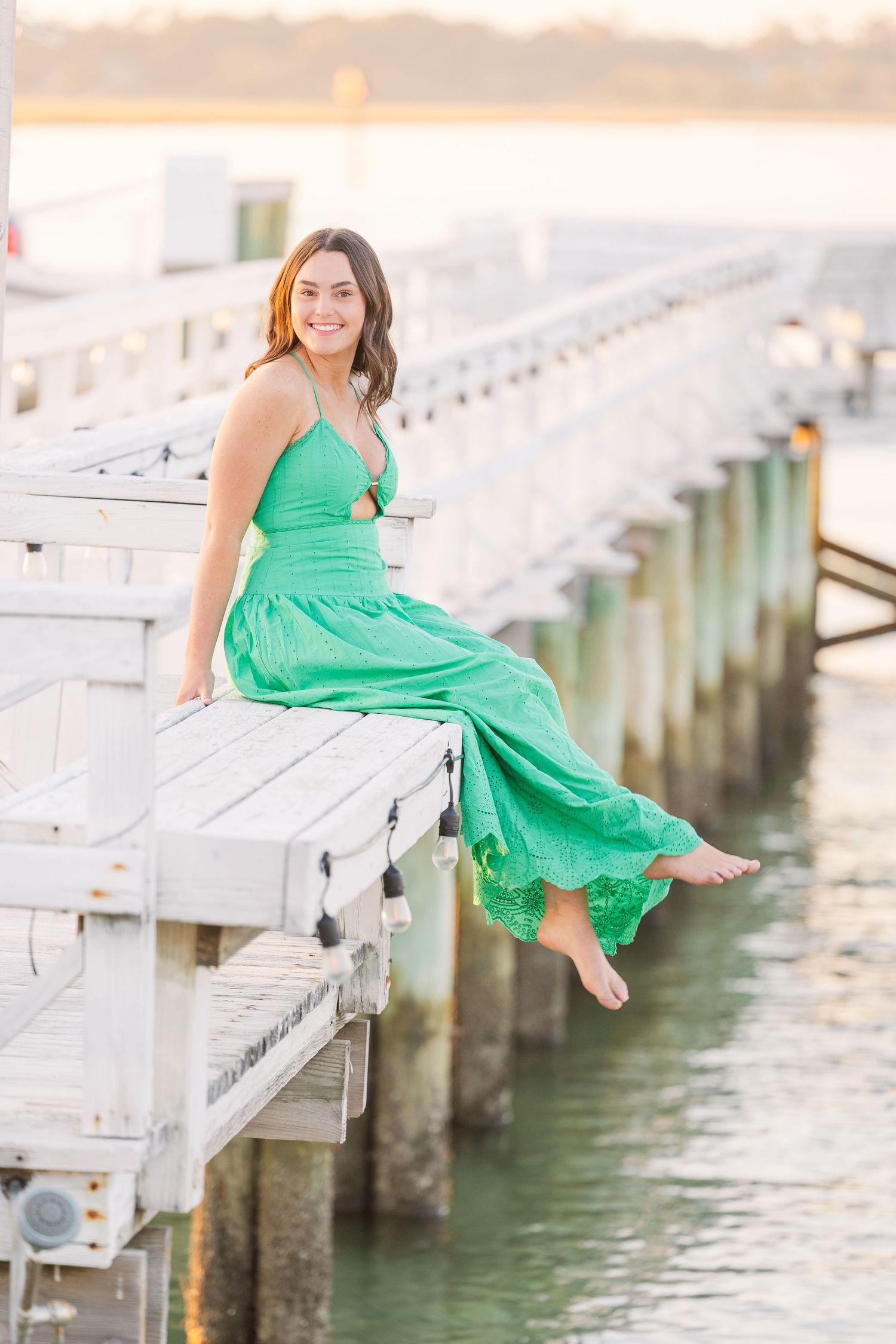 A green dress flows in the breeze while sitting on a white dock over calm waters.