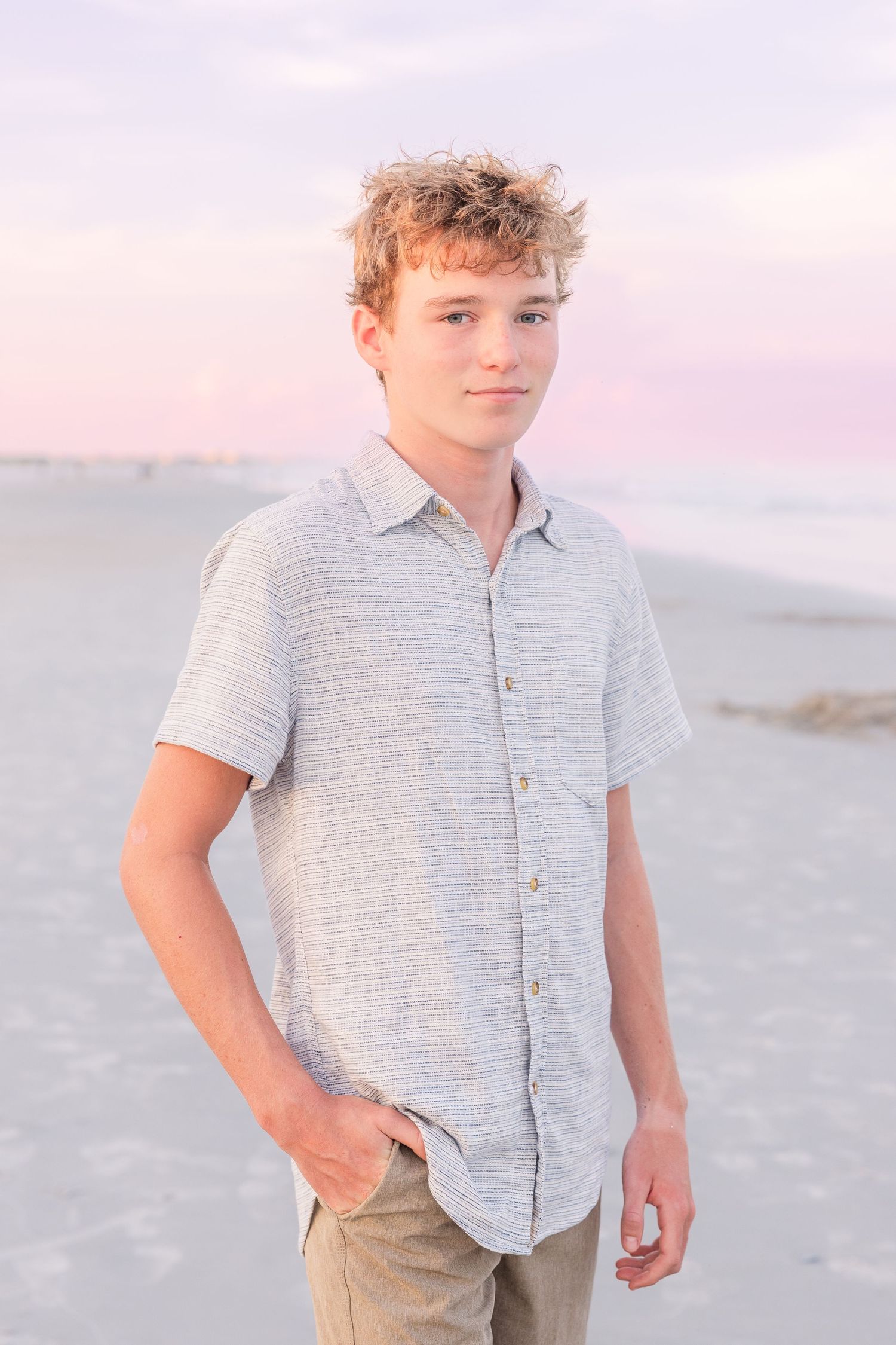 Boy in grey button-down shirt posing on beach during sunset.