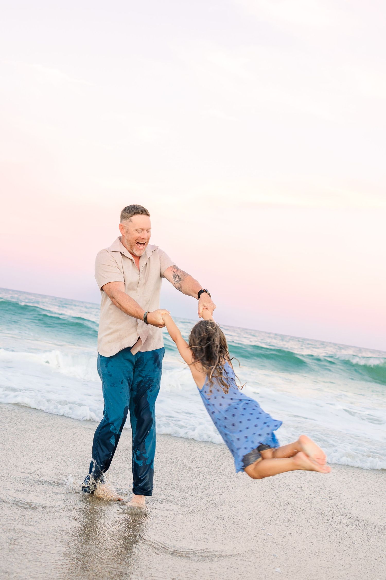 A laughing father swings his young daughter by the hands in the shallow ocean waves at sunset.