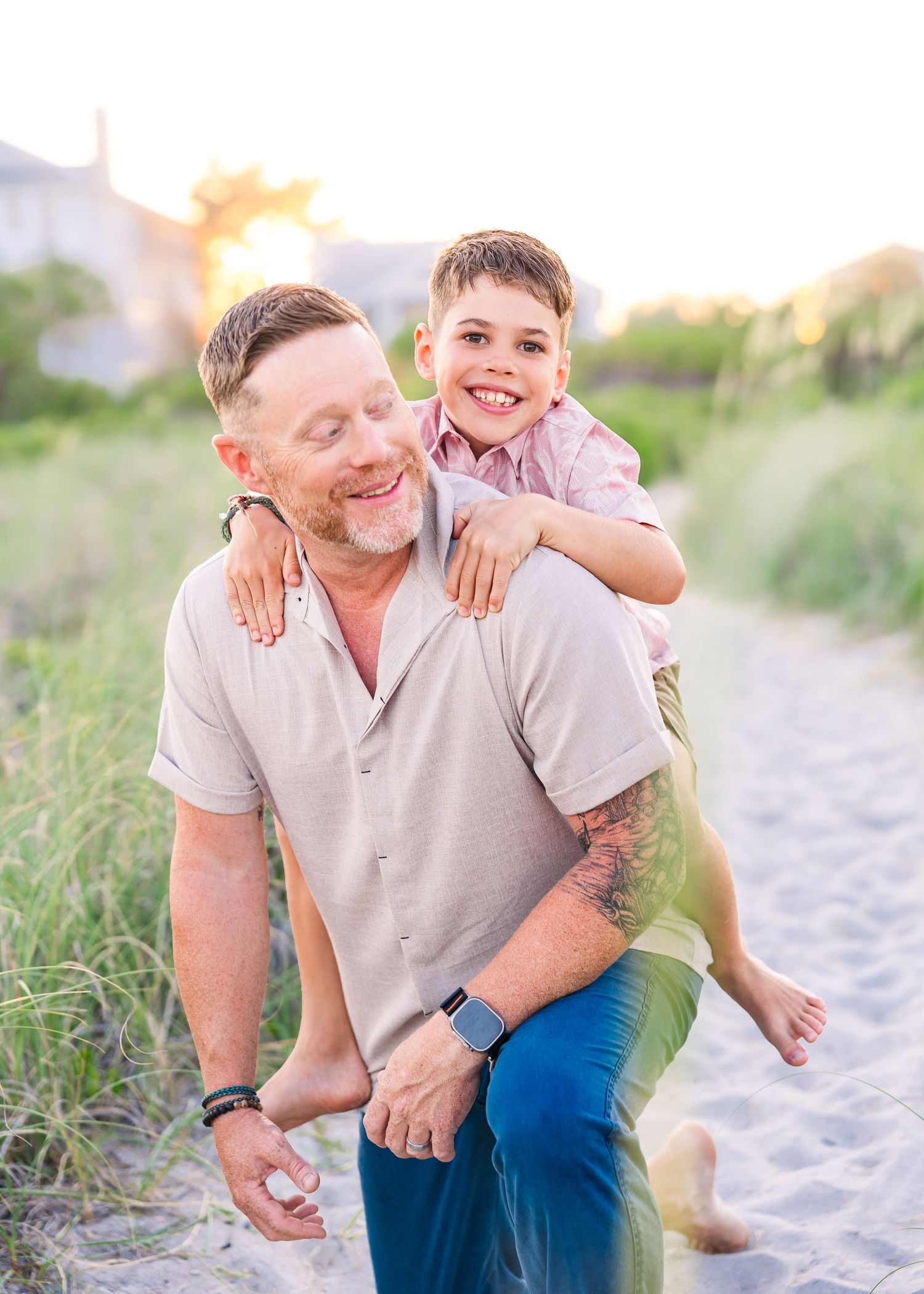 A man gives a smiling boy a piggyback ride on a sandy beach path at sunset.