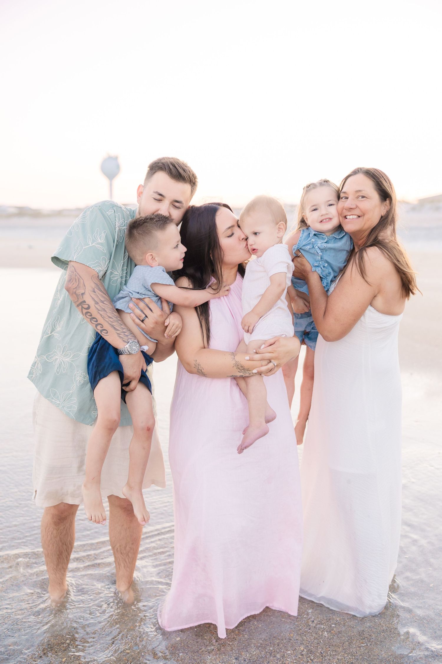 Family of five with three young children sharing kisses and hugs on a beach at sunset.