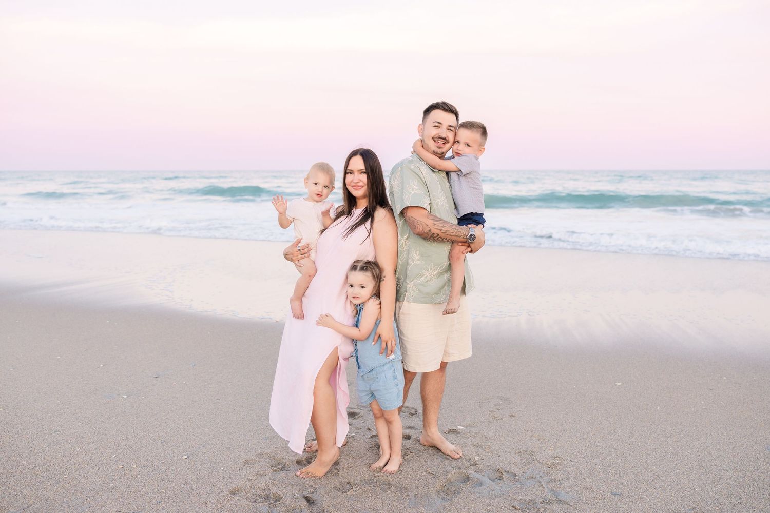 Family of five posing on a beach at sunset, parents holding children with ocean waves behind them.