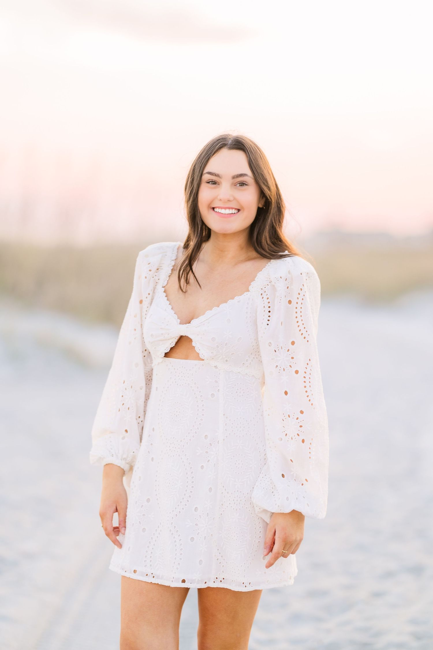 A white eyelet lace dress with balloon sleeves photographed on a beach during sunset.