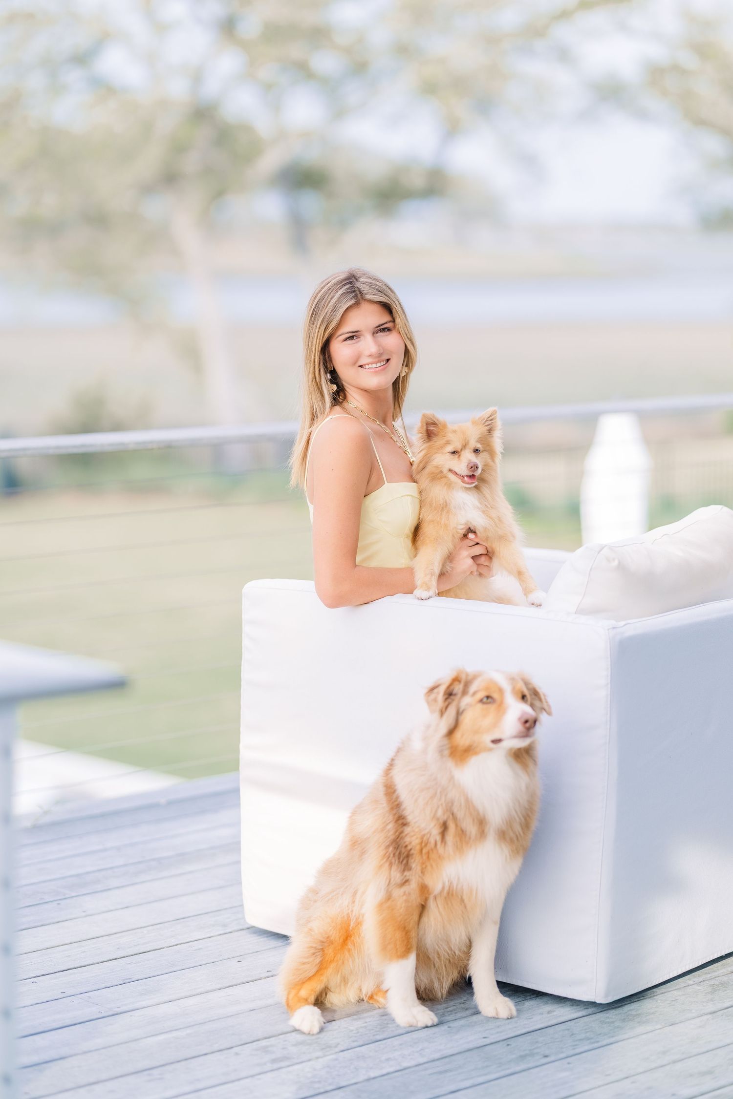 Two fluffy dogs pose with their owner on white furniture during an outdoor photoshoot.