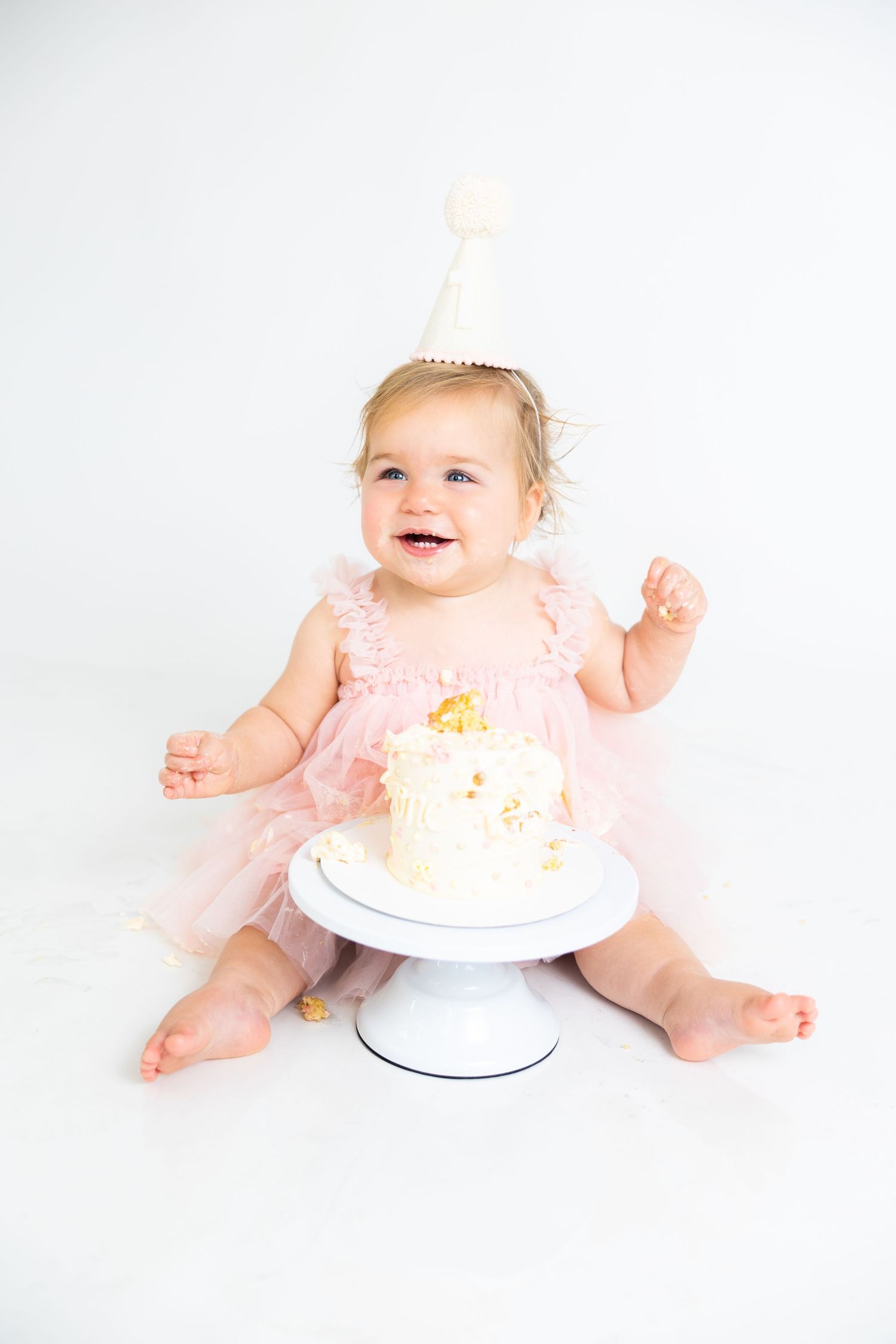 Baby in pink tulle dress and party hat smiling while sitting next to white birthday cake on cake stand.