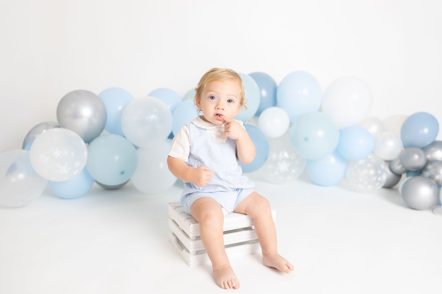 Baby in white outfit sitting on floor with decorative blue balloon arch.