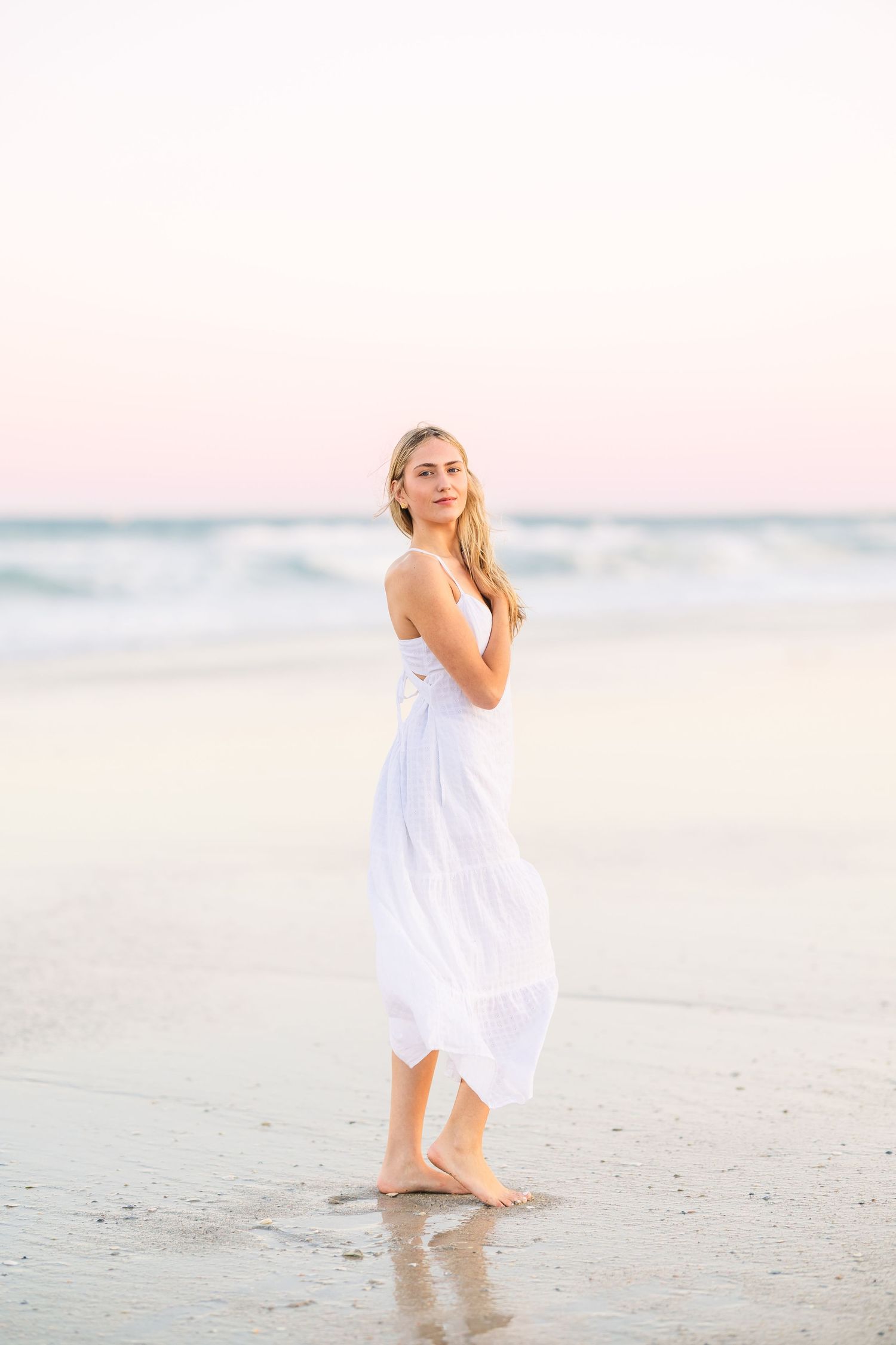 Serene beach portrait in flowing white dress at sunset with waves in background.