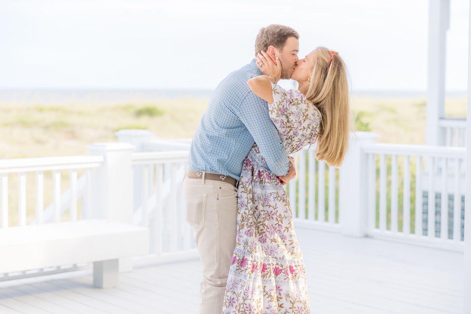 Romantic couple embracing and kissing on a beachfront porch in summer attire.