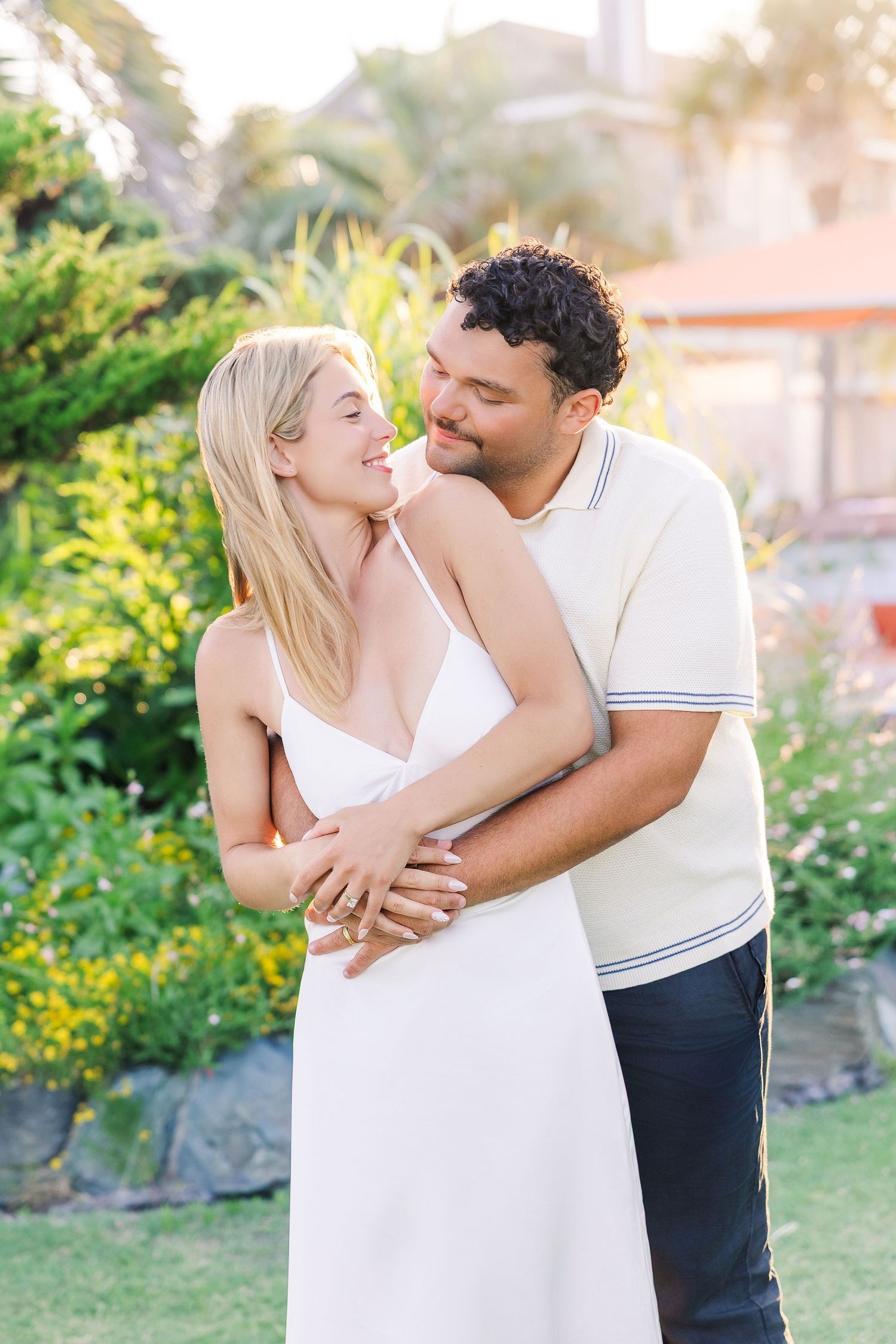 Couple embracing in a lush garden at golden hour, woman in white dress smiling up at man in cream polo shirt.