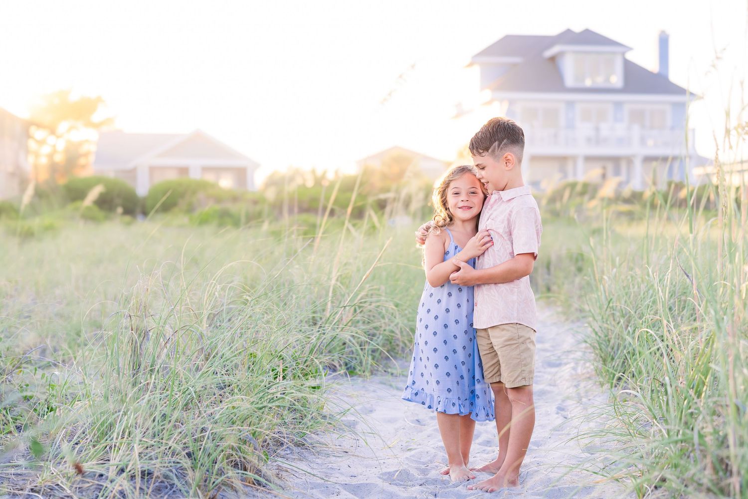 Two children embrace on a sandy beach path surrounded by tall grass at golden hour near a coastal home.