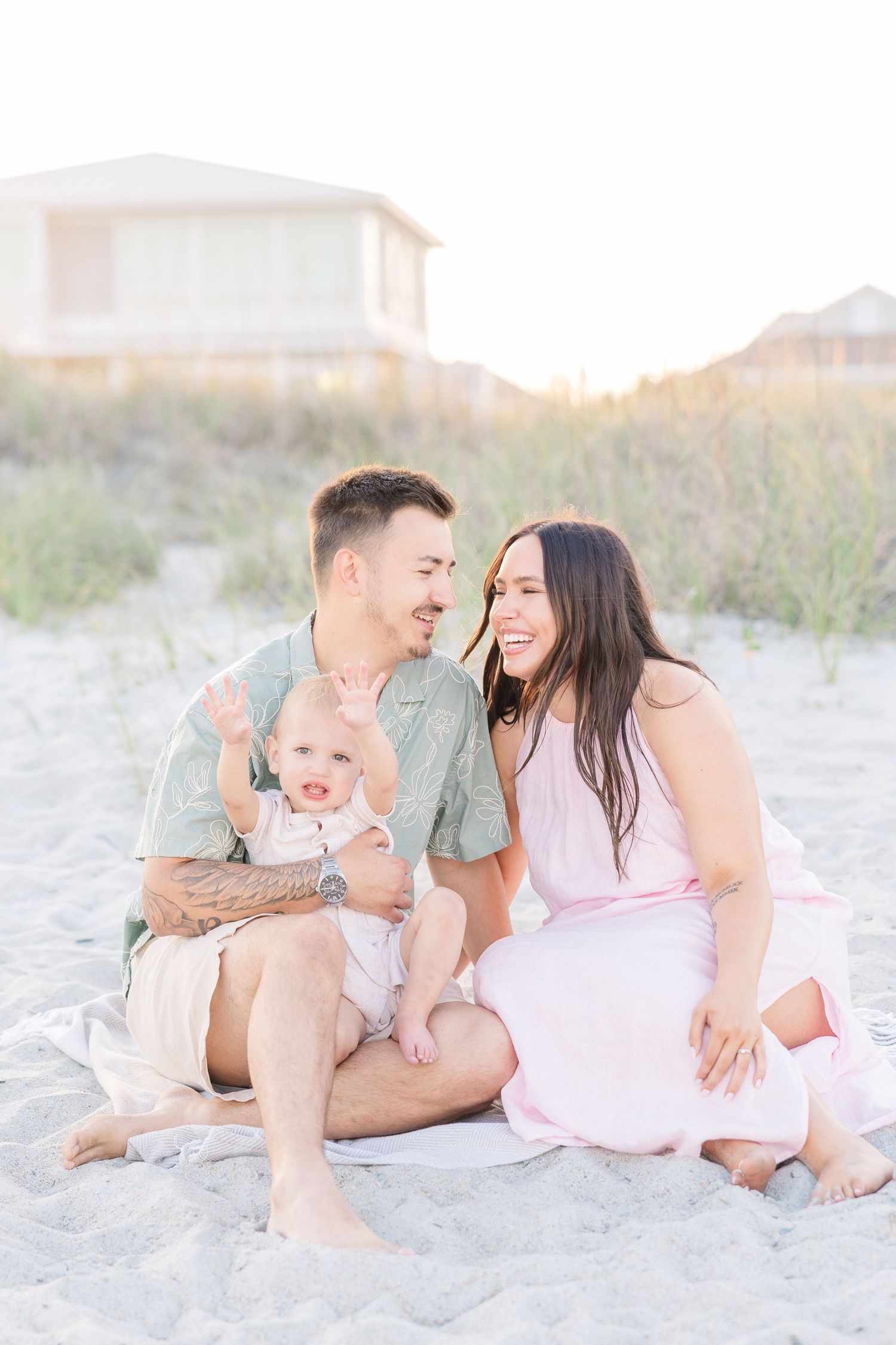 A couple sits on sandy beach laughing together while their baby waves at the camera.