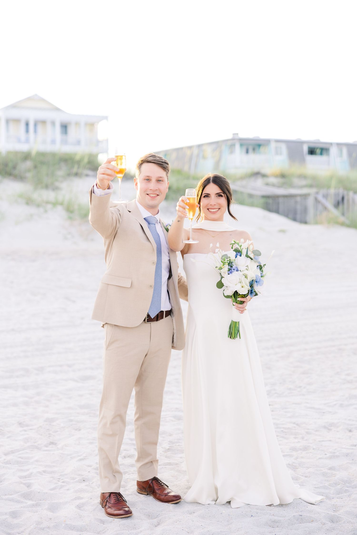 Bride and groom toast champagne glasses on sandy beach in tan suit and white gown in Carolina Beach near Wilmington, NC.