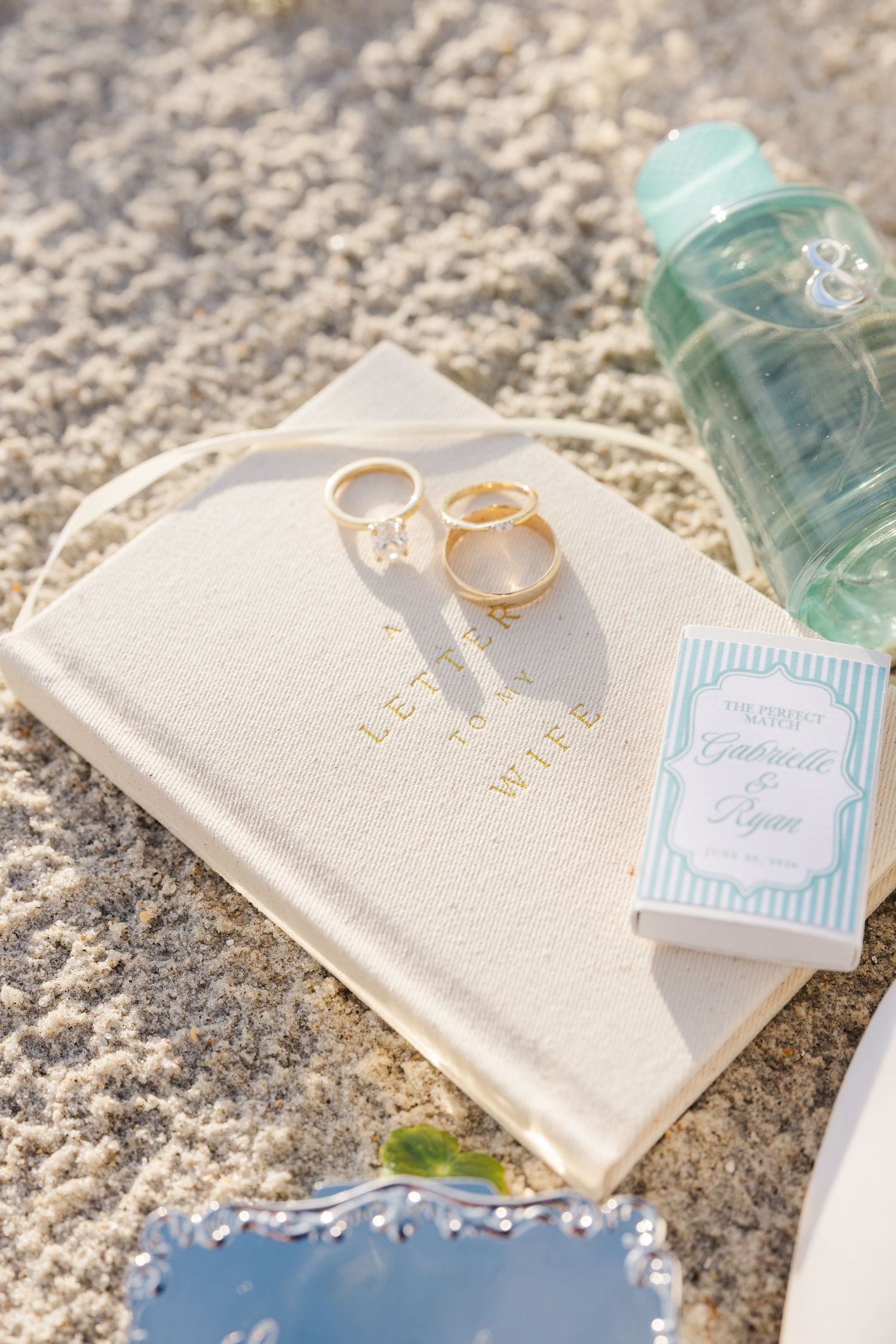 Gold wedding rings rest on a white leather journal beside a wedding program for Gabrielle and Ryan on sandy beach in Carolina Beach near Wil