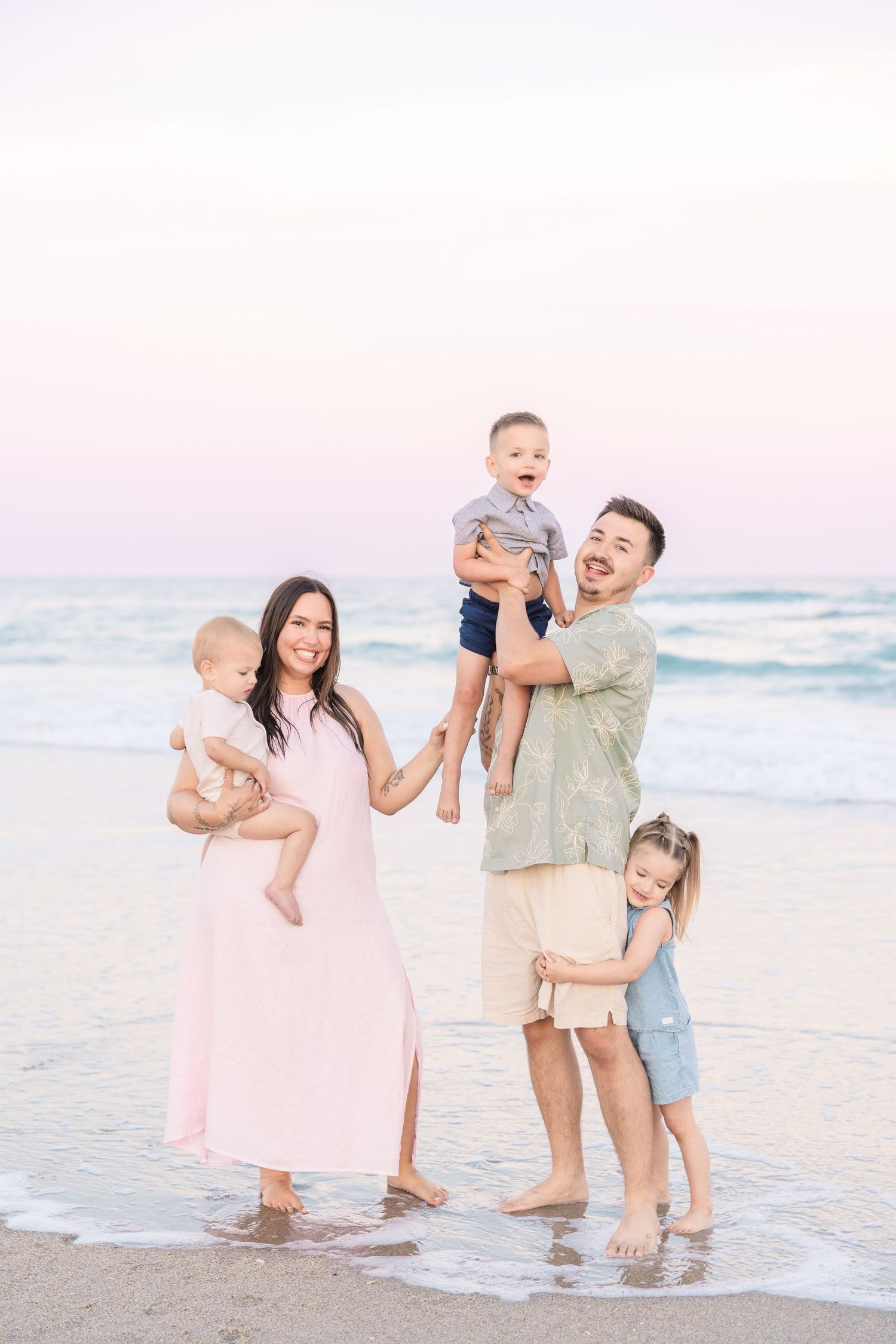 Joyful family of five posing at beach sunset, parents holding three young children near ocean waves.