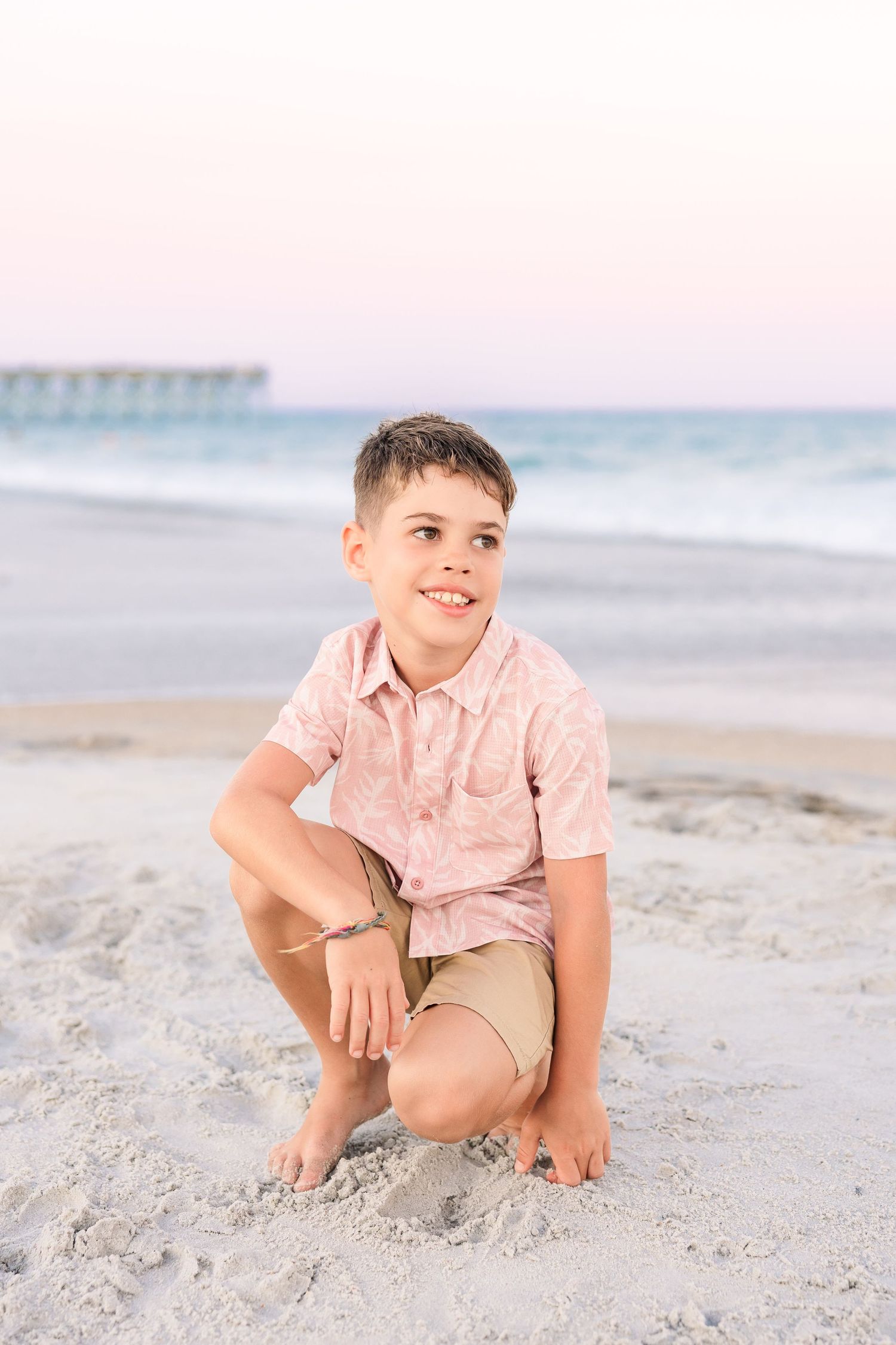 A smiling boy crouches on sandy beach wearing pink shirt and khaki shorts at sunset.