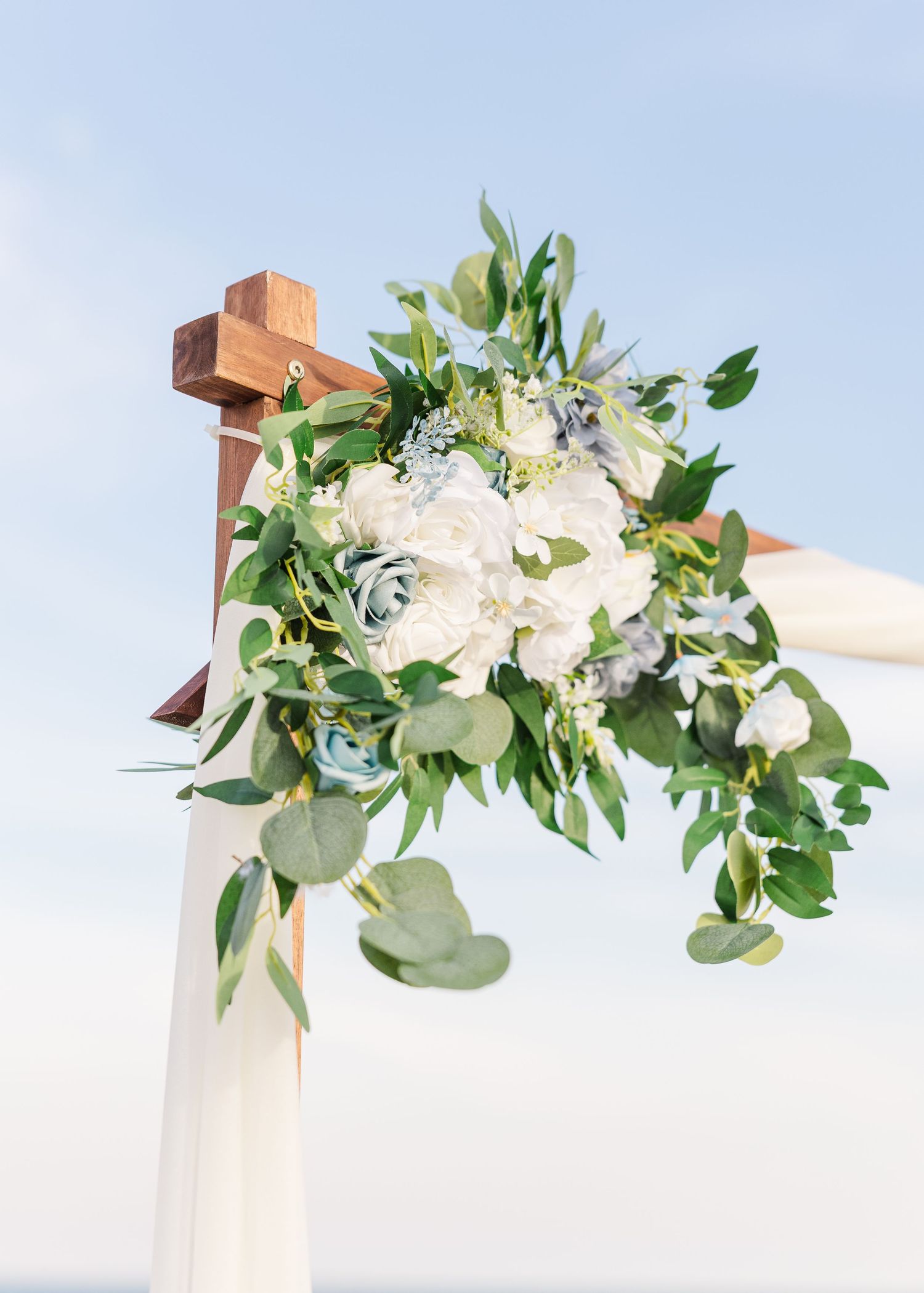 Wooden wedding arch adorned with white roses, blue flowers, and lush eucalyptus greenery against blue sky in Carolina Beach near Wilmington,