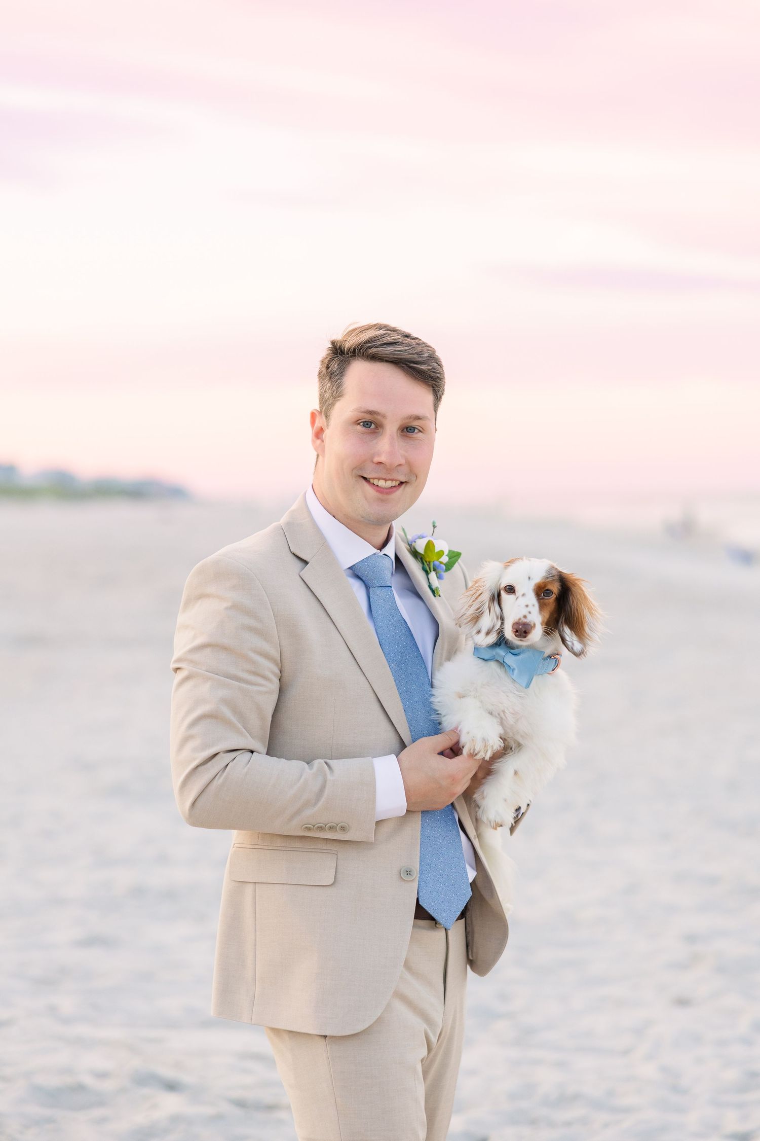 Groom in tan suit holds small dog wearing matching blue tie on beach at pink sunset in Carolina Beach near Wilmington, NC.