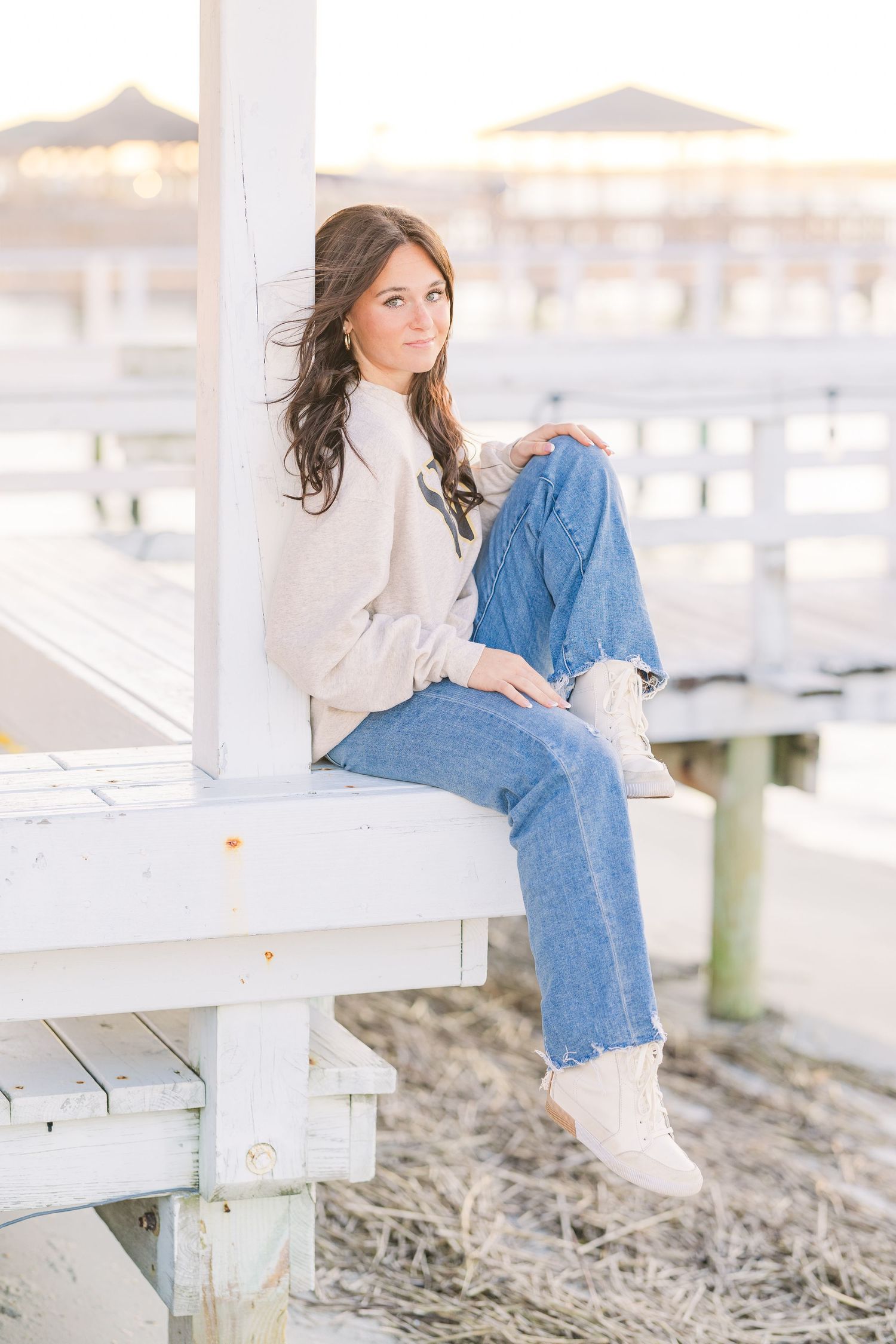 Casual beach portraits of someone in a white sweater and blue jeans sitting on a white wooden pier at sunset.