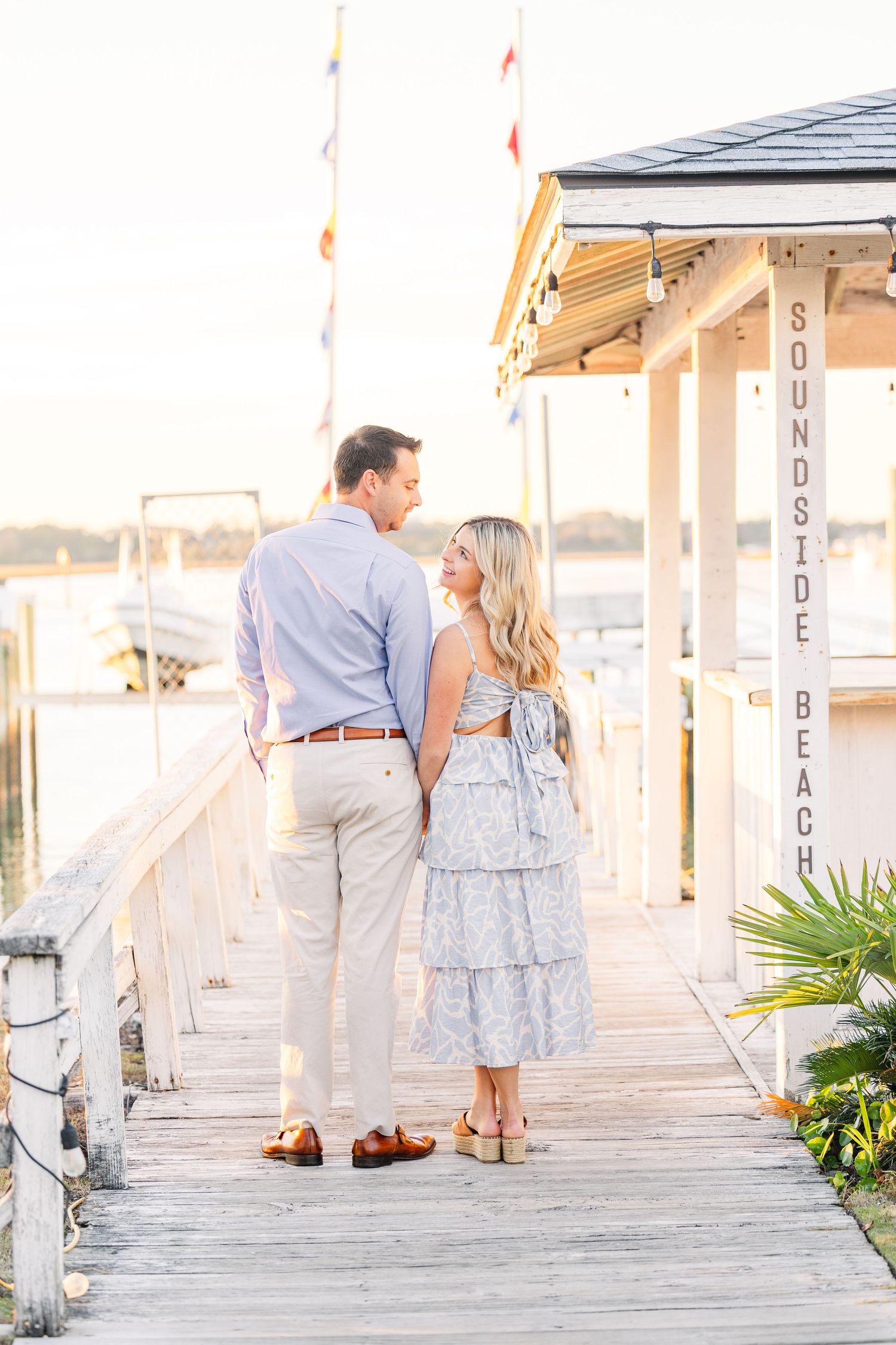 Couple in coordinated blue and white outfits walking along a waterfront dock at golden hour.