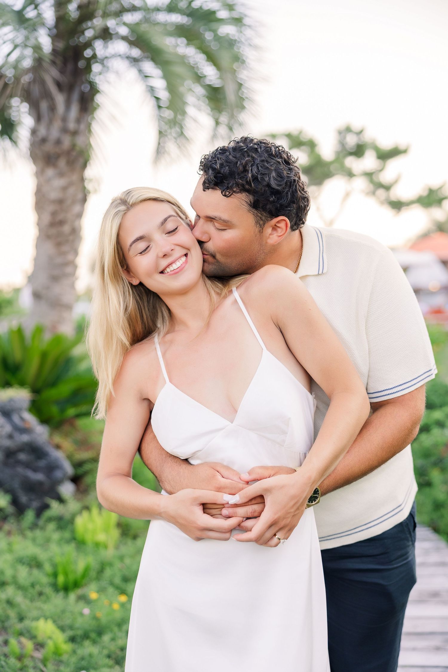 Man kissing smiling woman on the cheek while embracing her from behind in a tropical garden setting.