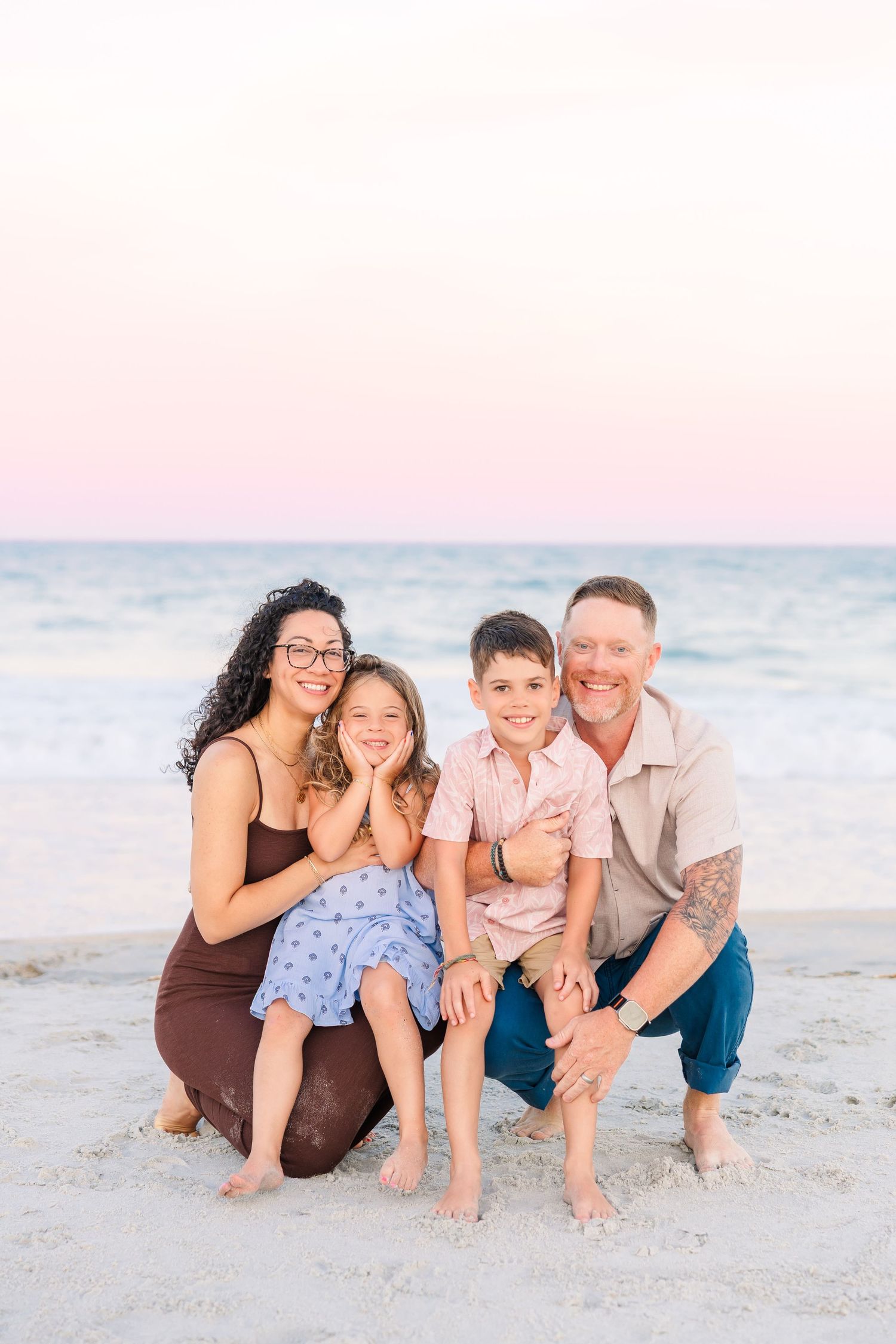 A happy family of four poses together on a sandy beach at sunset with ocean waves behind them.