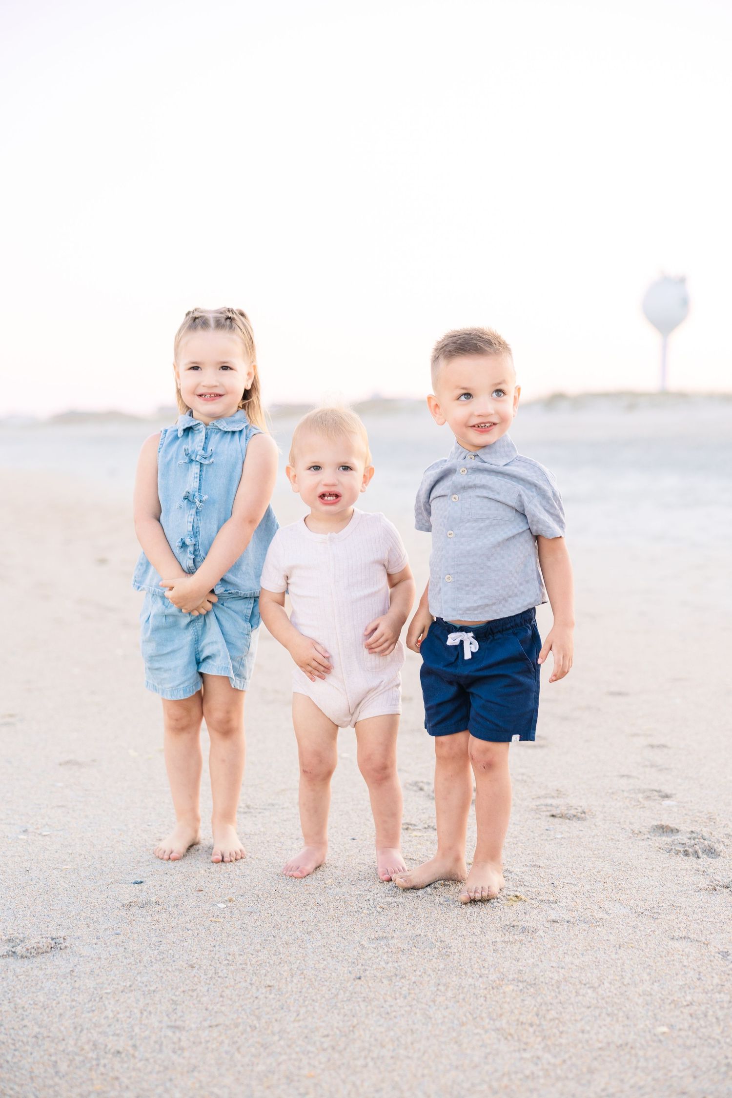 Three young children standing together barefoot on a sandy beach at sunset.