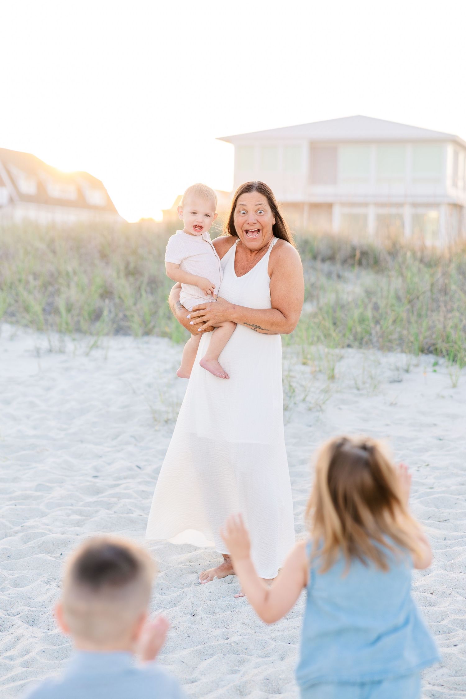 Grandmother in white dress holding baby on sandy beach dunes at golden hour while children run toward her.