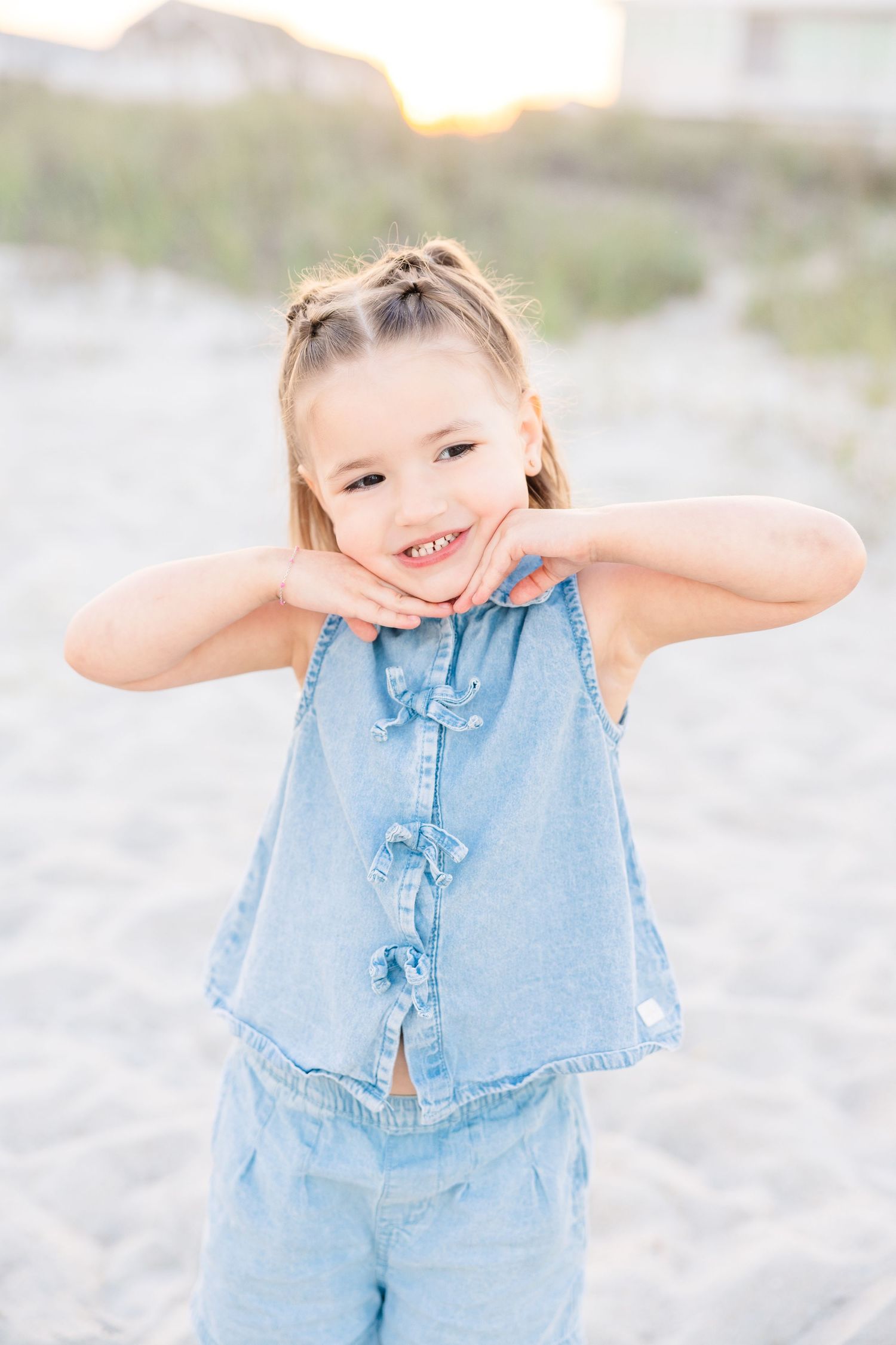 A smiling little girl in a denim romper poses playfully on a sandy beach at sunset.