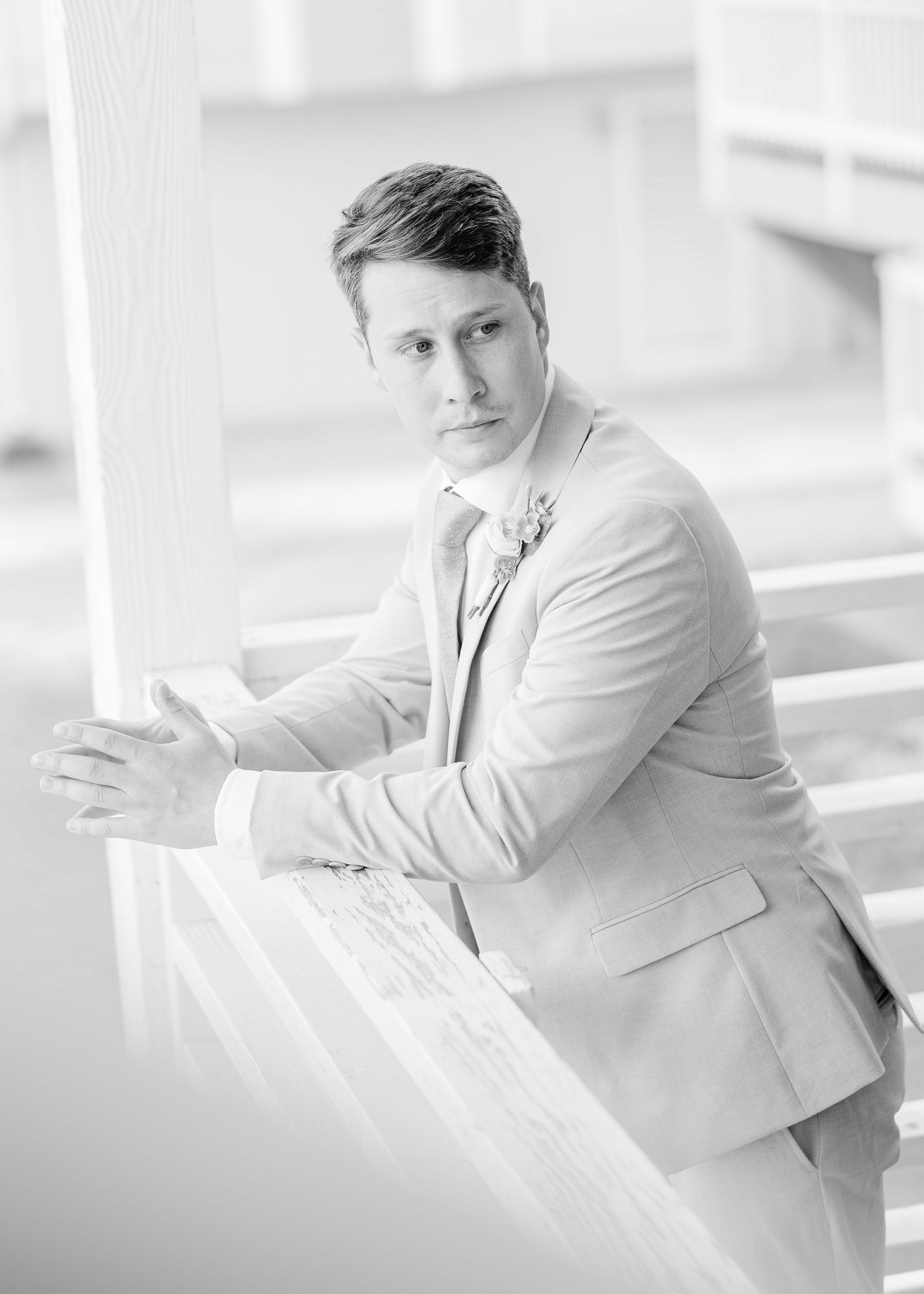 Black and white portrait of a dapper groom in a light suit with boutonniere leaning on white railing in Carolina Beach near Wilmington, NC.