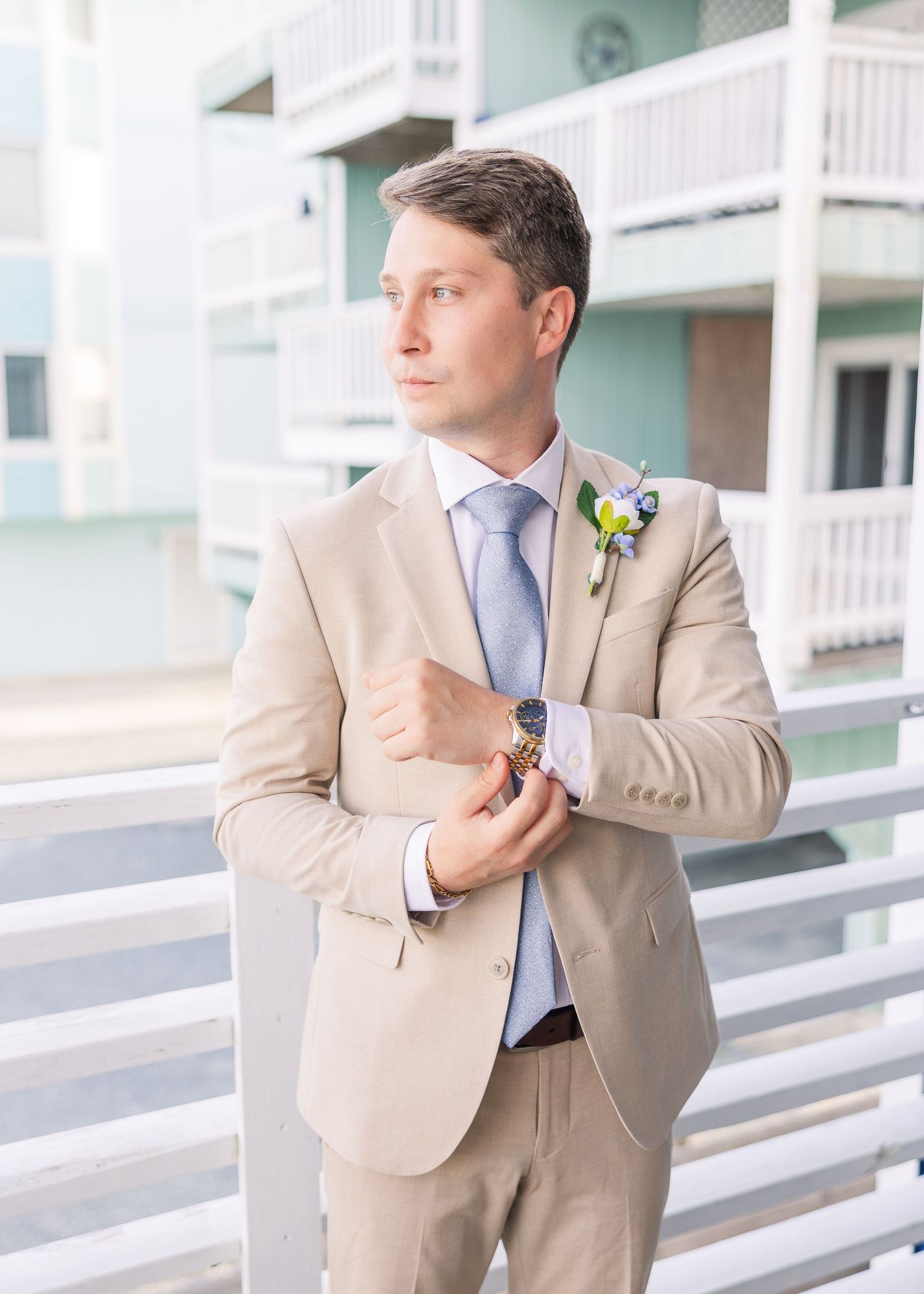 Groom in tan suit with blue tie and boutonniere adjusting cufflinks on a balcony at a beach wedding.
