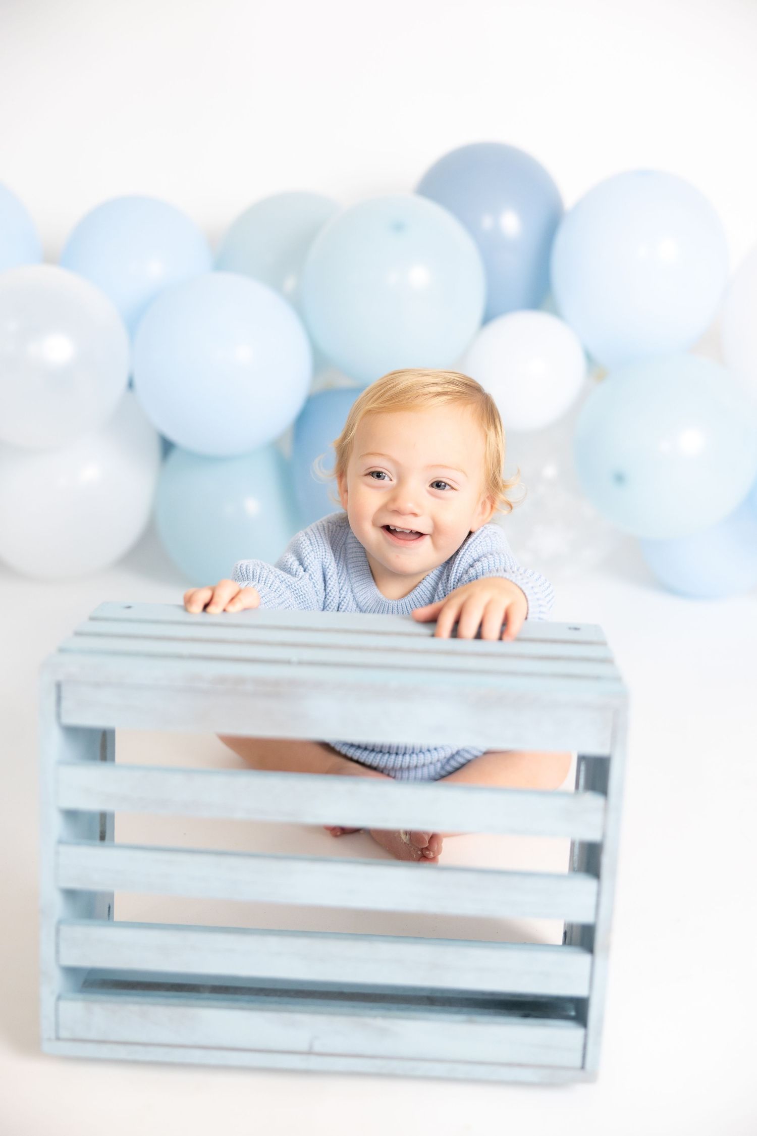 Smiling baby plays with blue wooden crate against balloon backdrop.