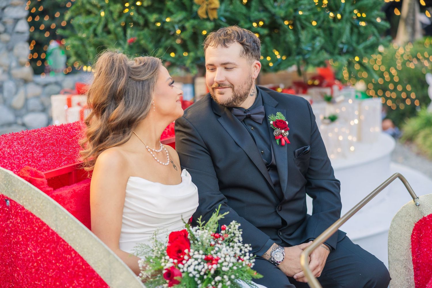 A couple sitting together on a red bench surrounded by greenery and twinkling lights.
