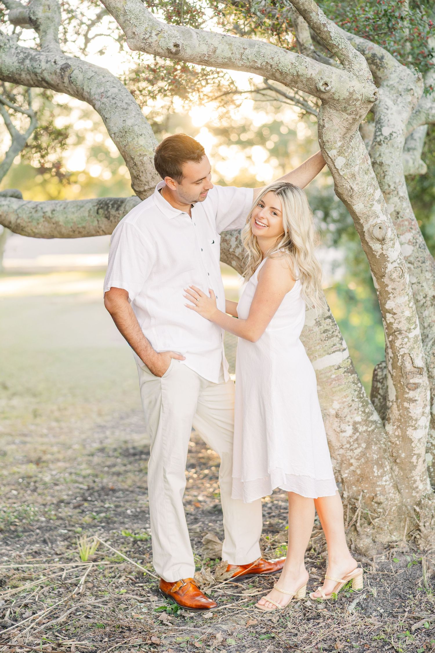 Couple in white clothing sharing romantic moments under large tree branches at sunset.
