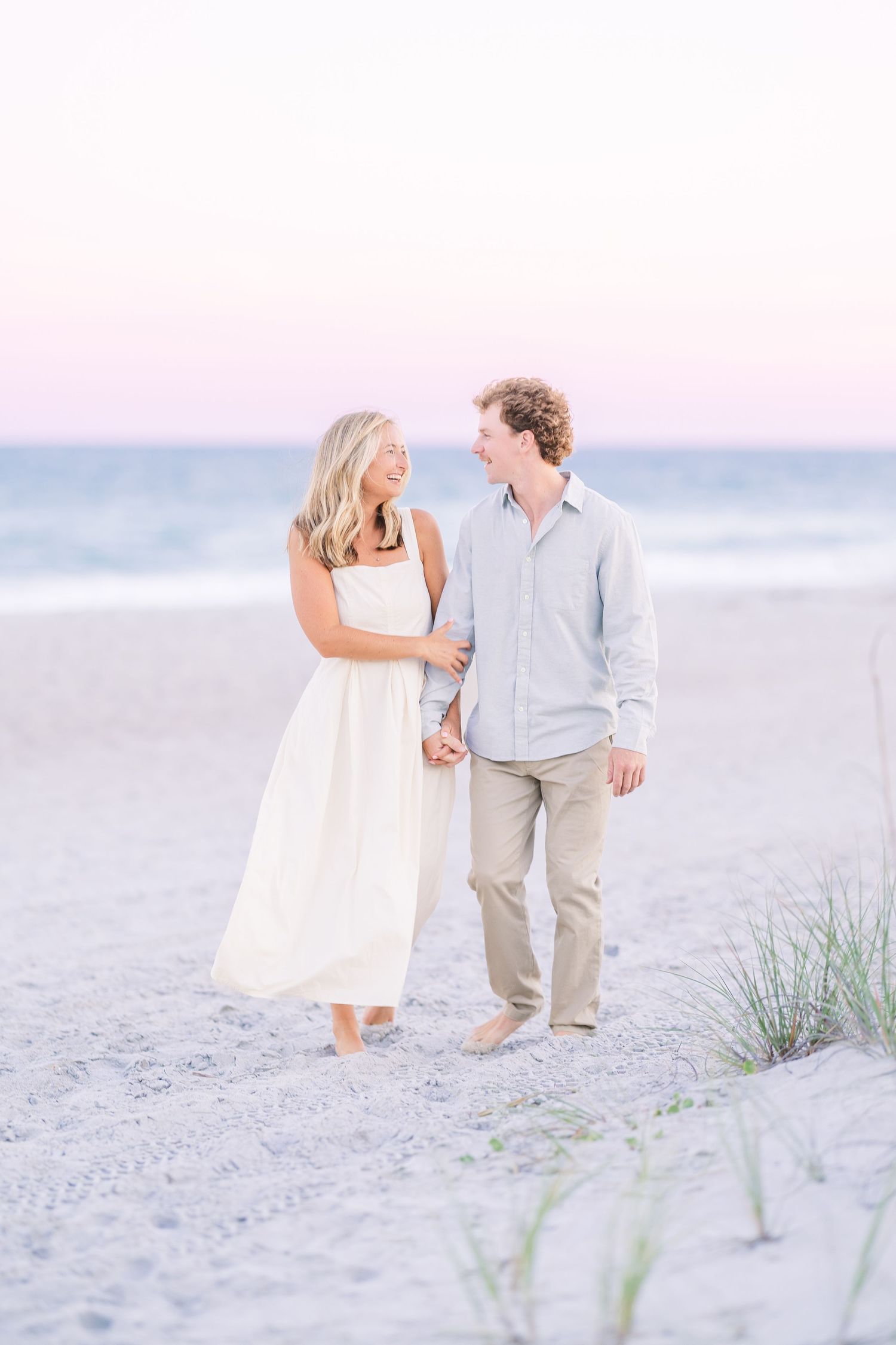 Couple walking barefoot along serene beach landscape during pastel sunset.