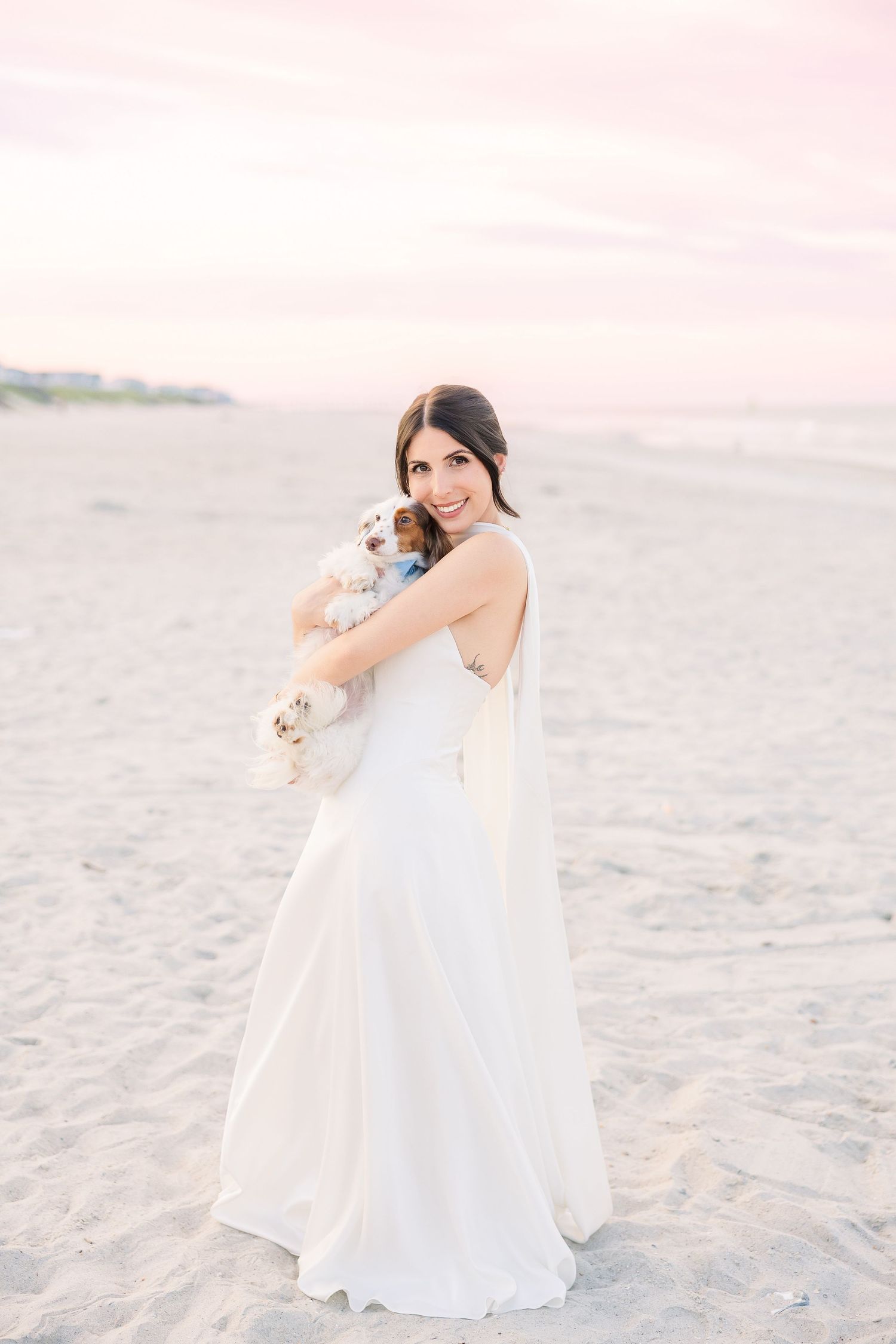 Radiant bride in white gown holds a small dog on a sandy beach at sunset with pink sky in Carolina Beach near Wilmington, NC.
