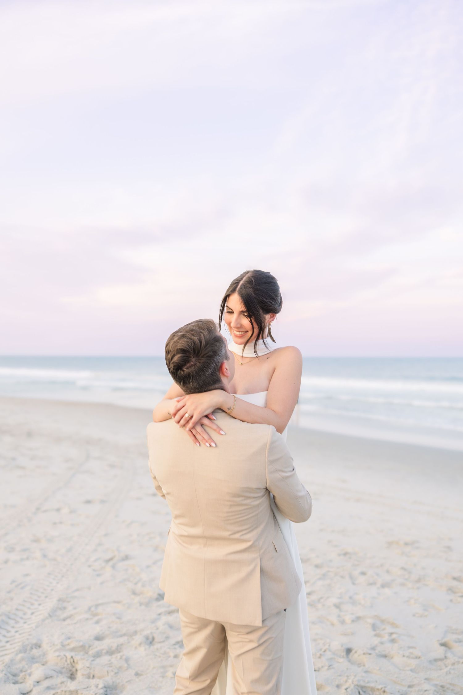 Bride and groom embrace on a sandy beach at sunset, woman in white gown smiling at man in tan suit in Carolina Beach near Wilmington, NC.