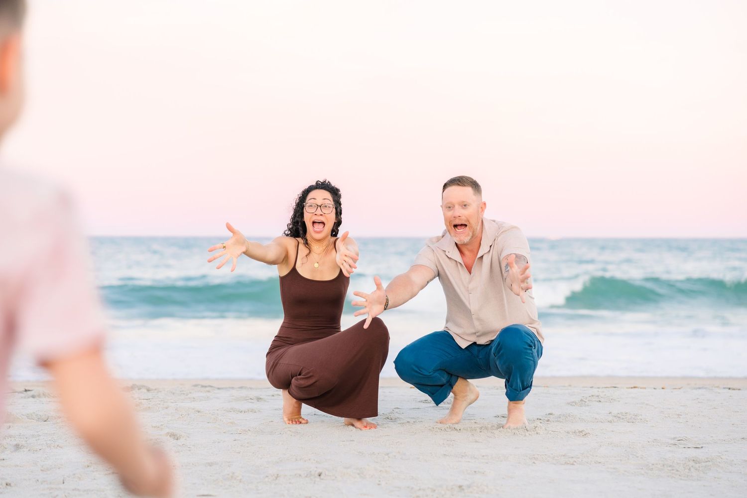 Excited couple crouching on a sandy beach with arms outstretched, greeting someone near the ocean at sunset.