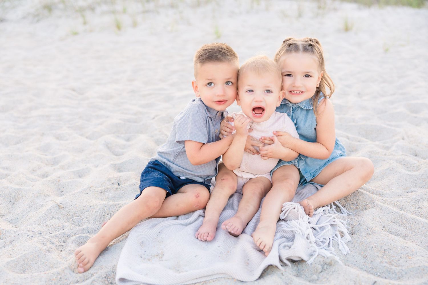 Three young siblings sitting together on a blanket on a sandy beach.
